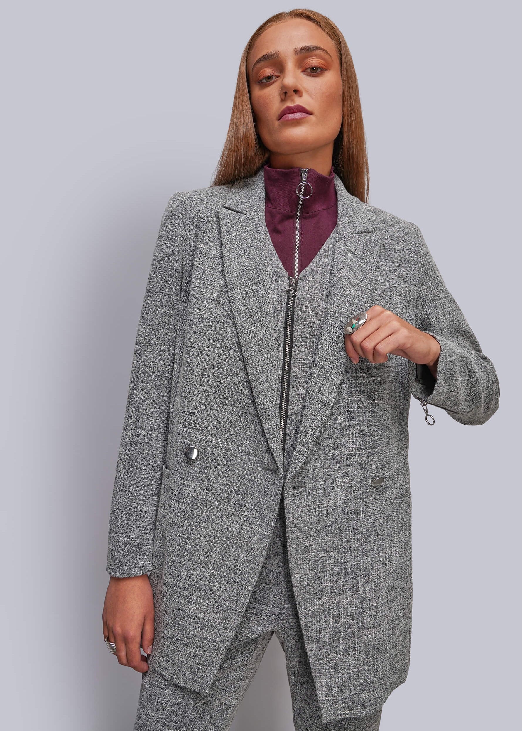 A woman modeling a gray textured blazer and matching trousers with a zippered waist, worn over a dark purple high-neck top, against a solid light gray background.