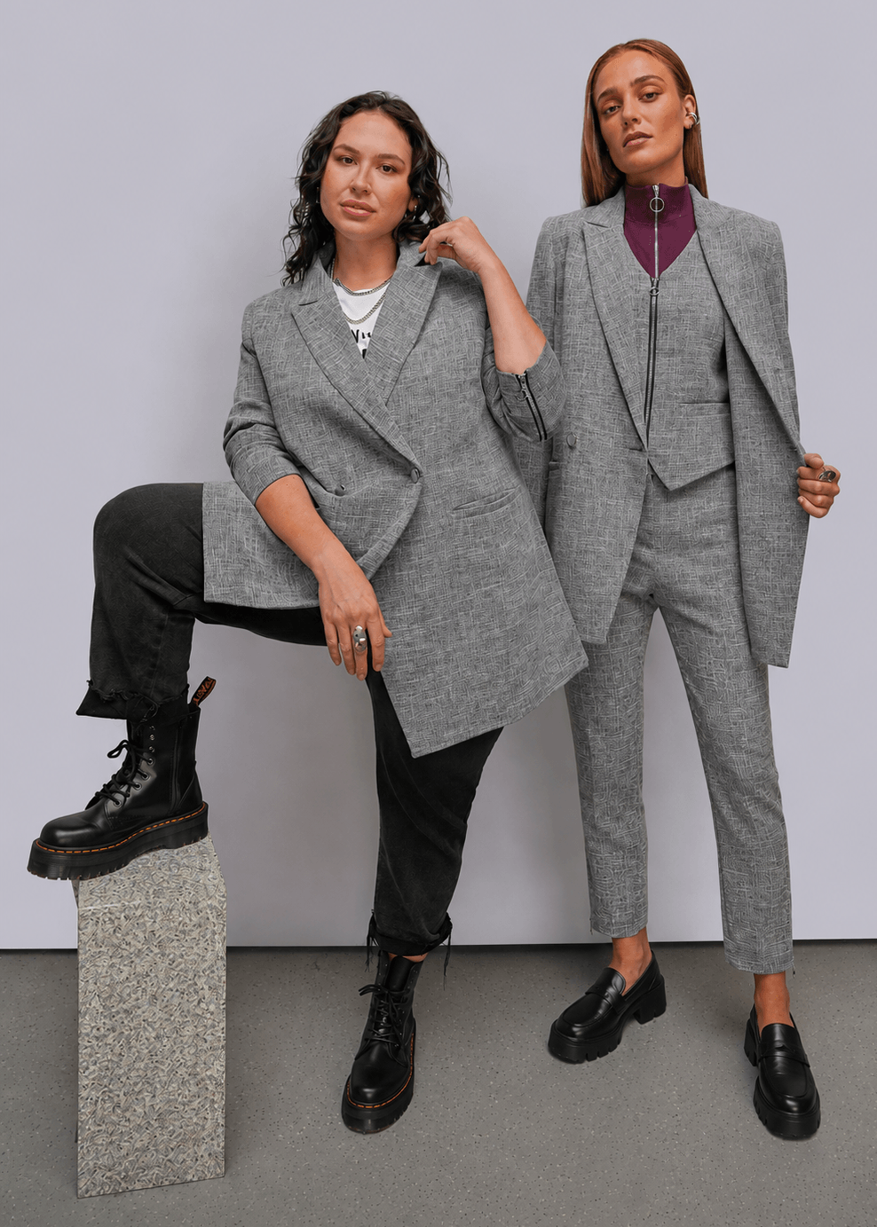 Two models posing in gray tailored suits against a light gray studio background. The model on the left wears an oversized gray blazer with black pants and boots, sitting on a stone block.
