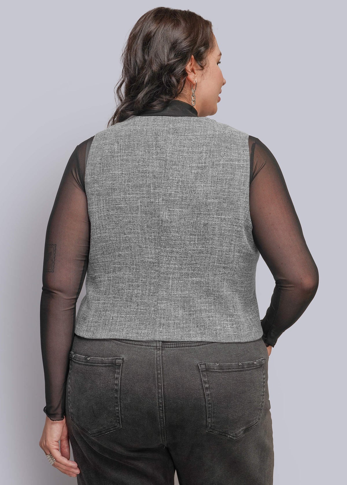 1X=A person shown from the back wearing a gray textured vest over sheer black long-sleeve mesh top, paired with dark gray jeans, against a solid light gray background.