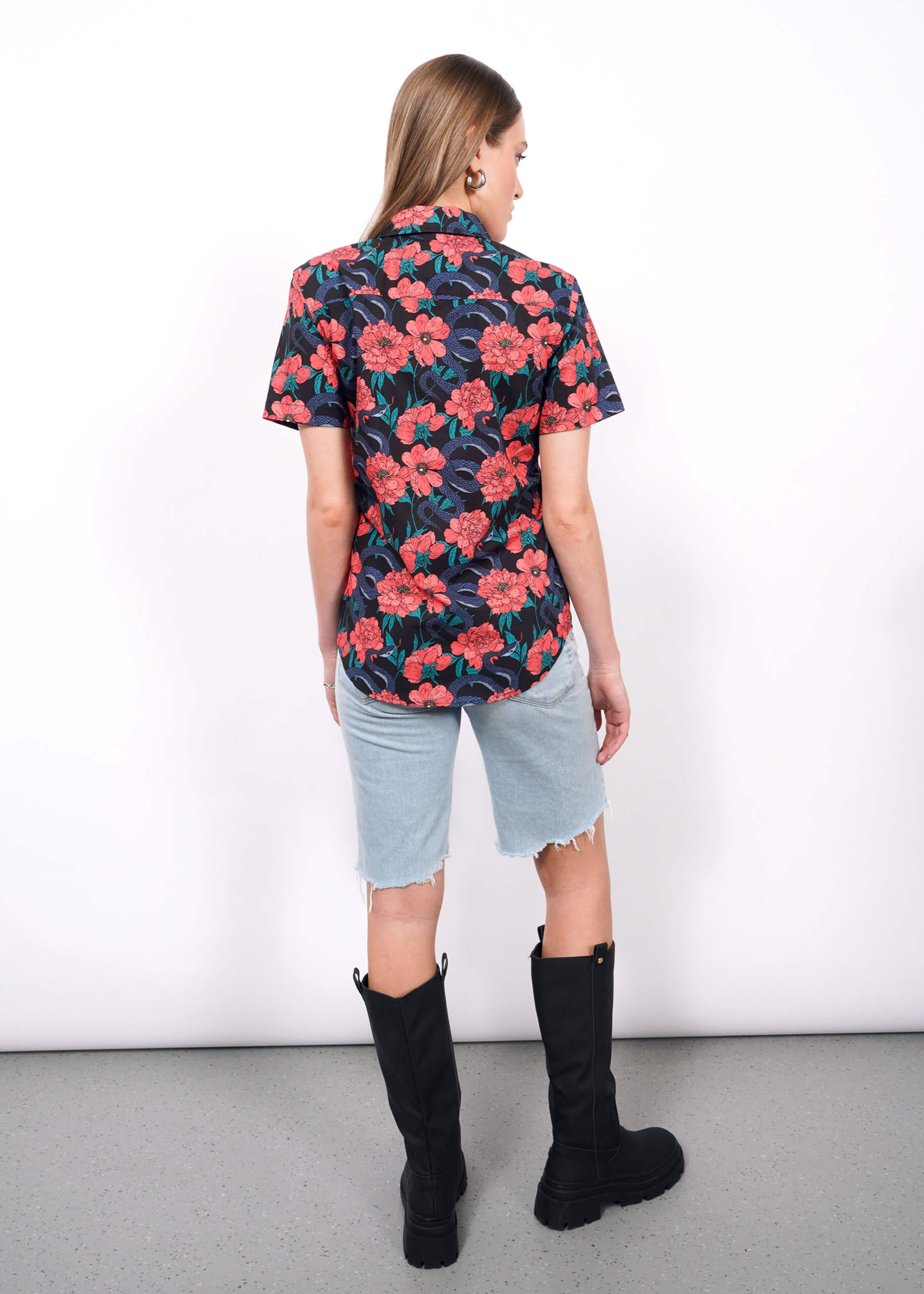 A woman stands facing away, wearing The Essential Button Up in tailored navy with a red floral pattern, light denim knee-length shorts, and black knee-high boots. She stands on a gray floor against a plain white wall.