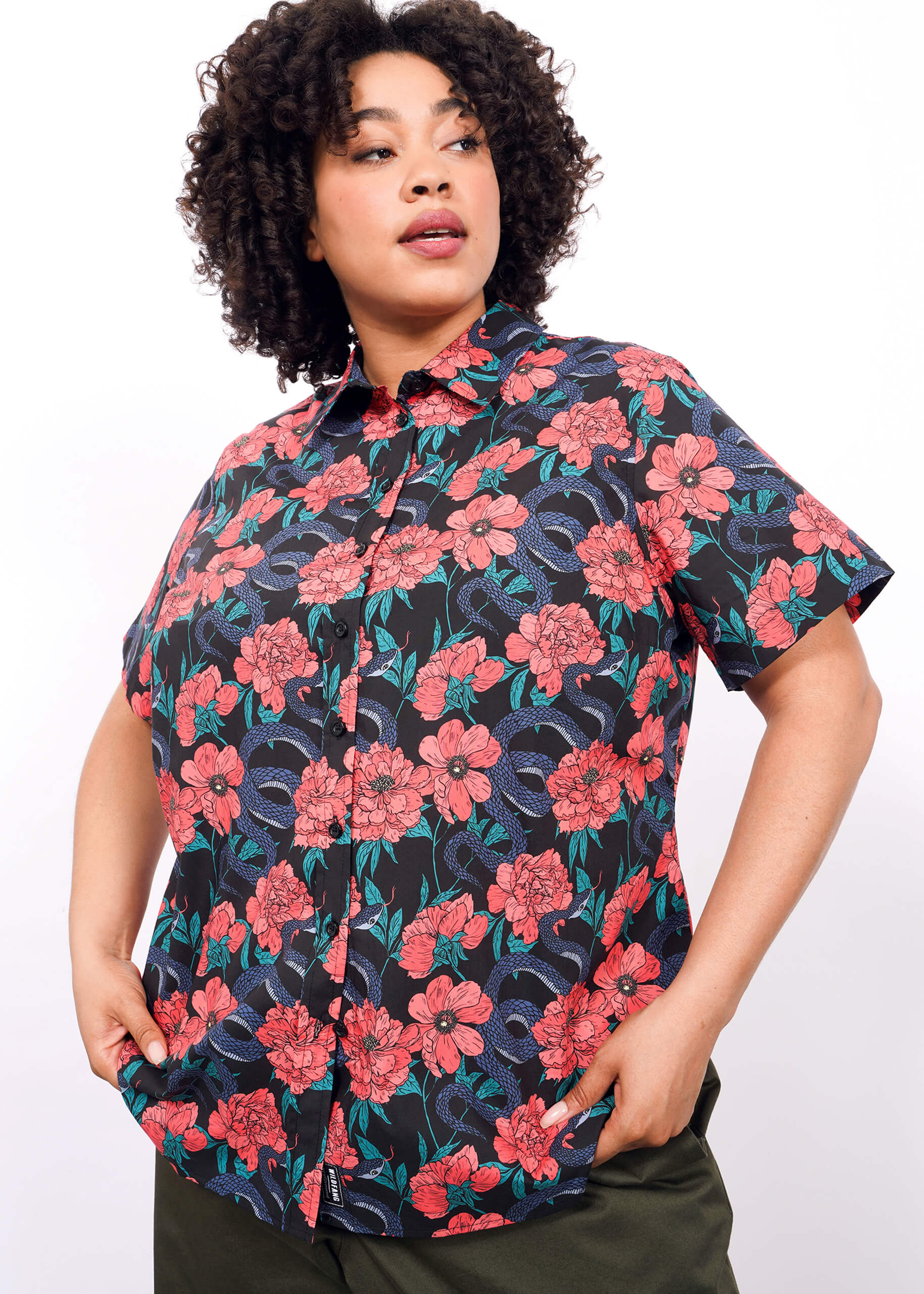 A person with curly hair wears The Essential Button Up, a bright red and purple floral cotton shirt with a tailored fit, paired with dark green pants, posed against a plain white backdrop.
