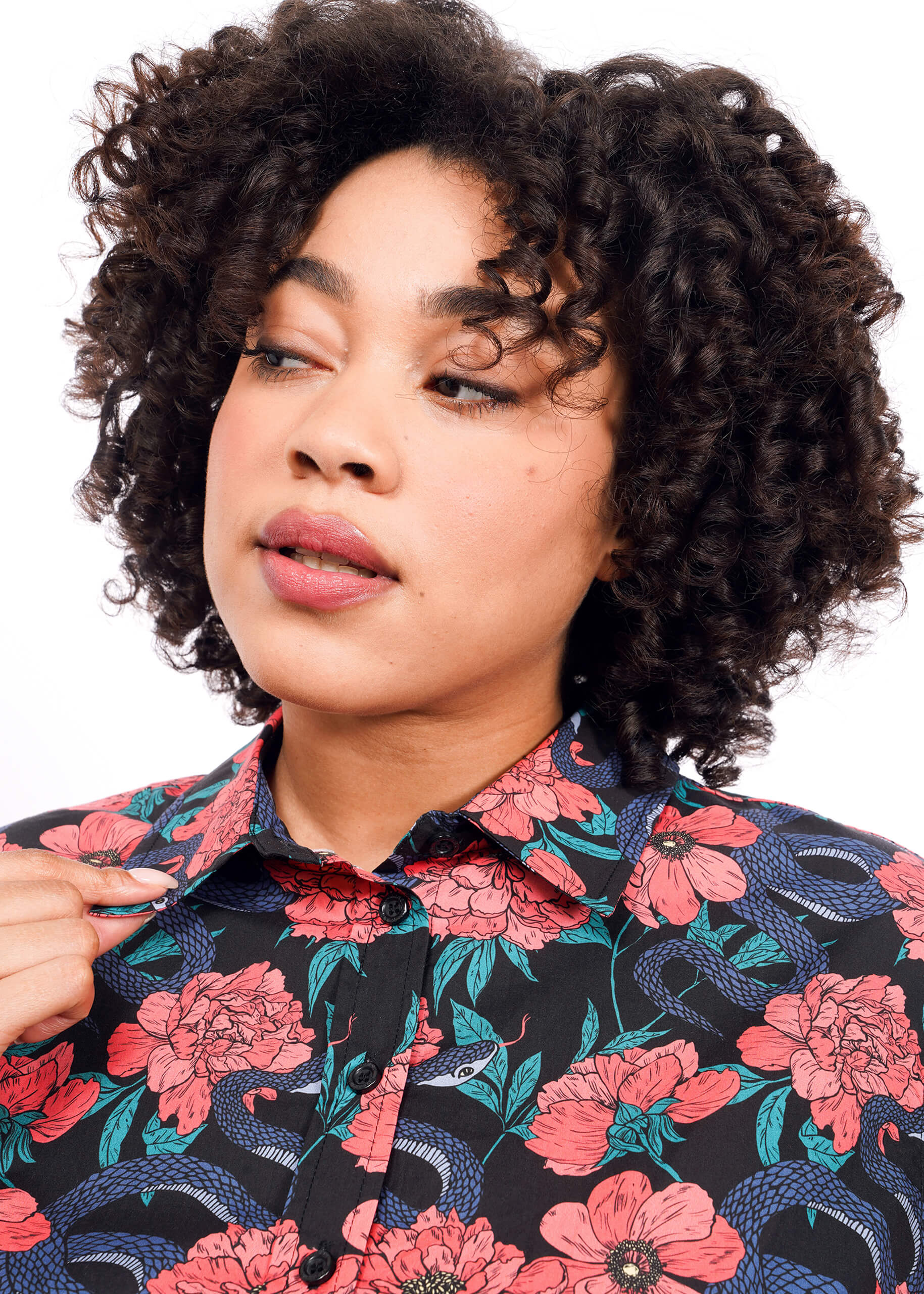 A person with curly hair looks to the side, wearing The Essential Button Up—a tailored fit black shirt with pink flowers and blue snakes.