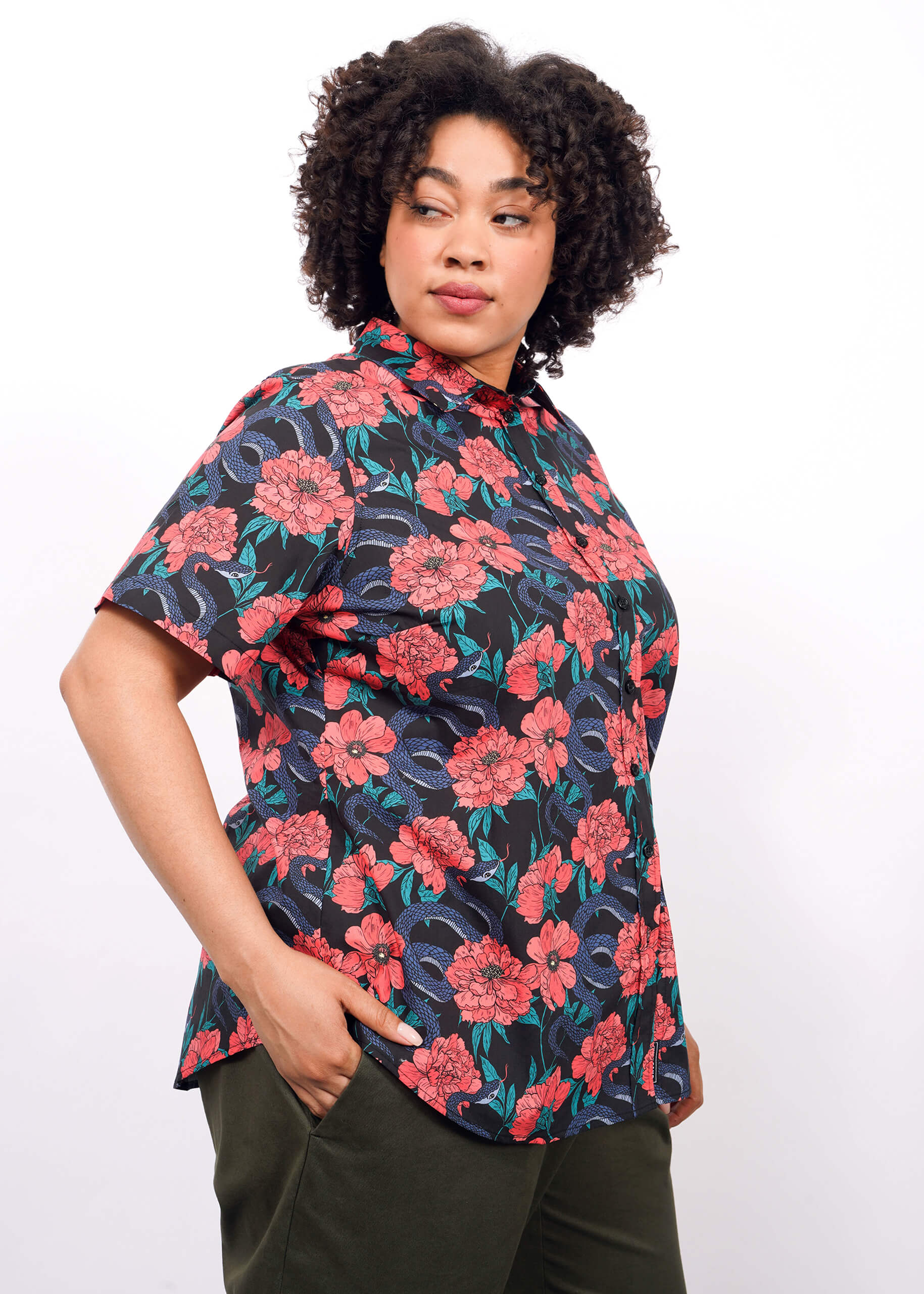 A person with curly hair wears The Essential Button Up, tailored fit, short-sleeve, in a vibrant red and blue floral pattern, paired with dark green pants. They stand against a plain white background, looking to the side with hands in their pockets.