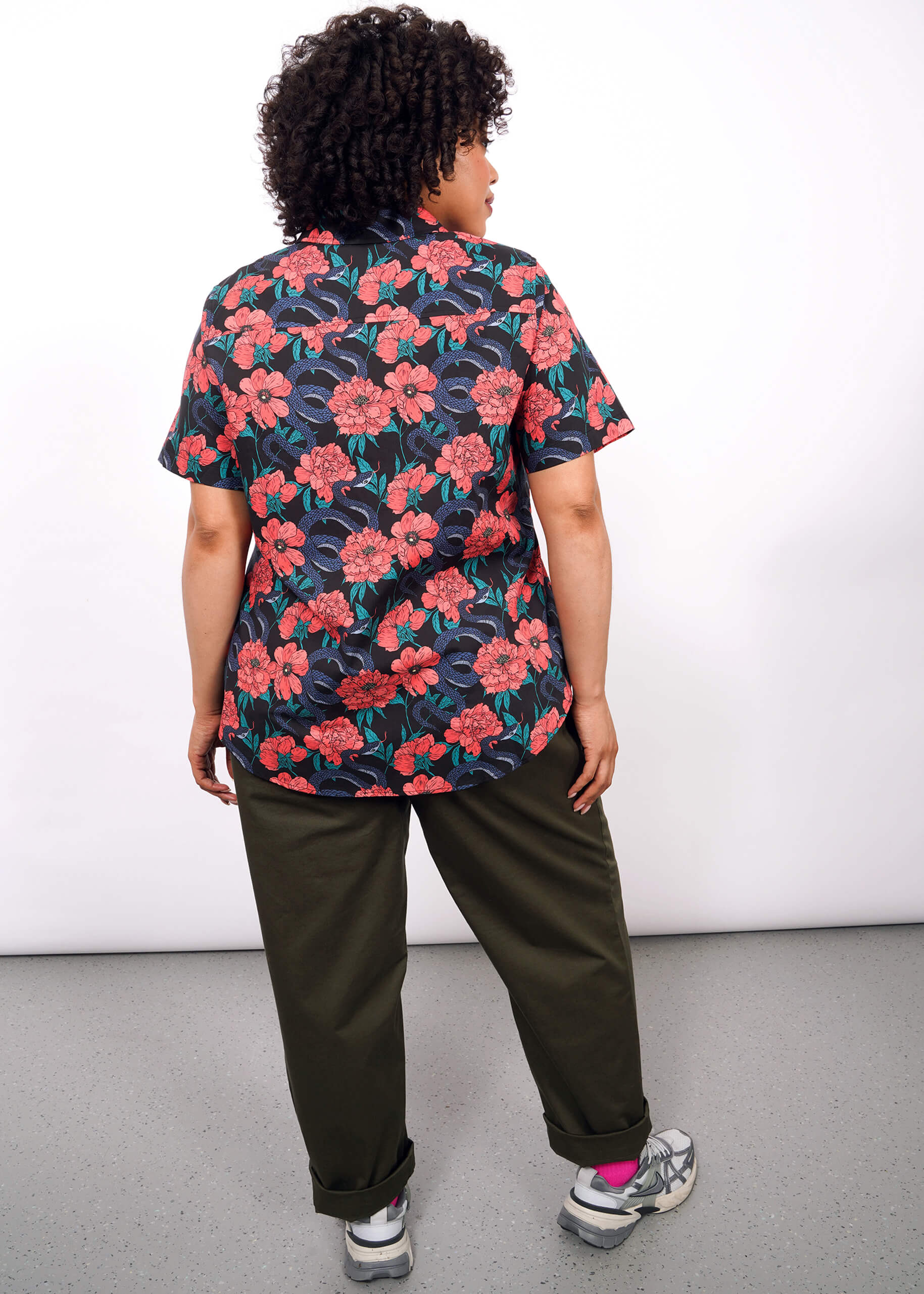 A person with curly hair stands facing away, wearing The Essential Button Up—a bold red floral print short-sleeve shirt in breathable cotton—paired with olive green pants and sneakers. The background is plain white.