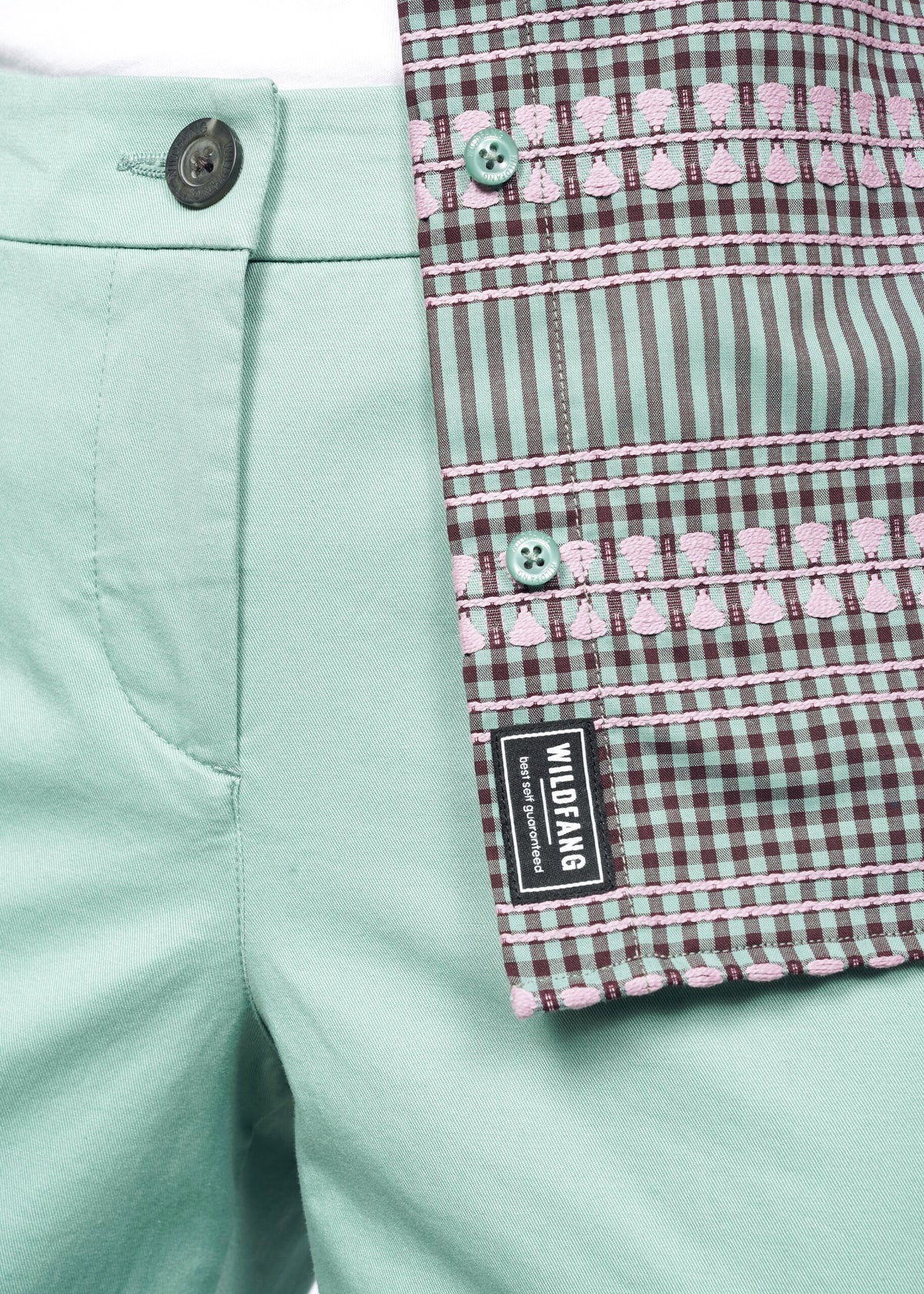 Close-up image of the Essential Button Up in Gingham Sage, with sage Trousers, showing the Wildfang - best self guaranteed - label 