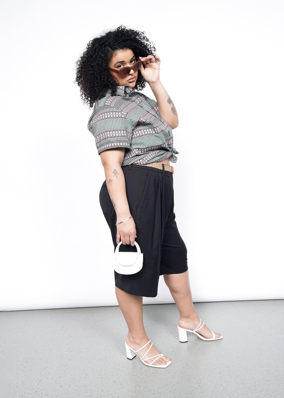 Side image of model wearing the Essential Button Up in Gingham Sage in size 1X, with black shorts, a white handbag and white strappy shoes