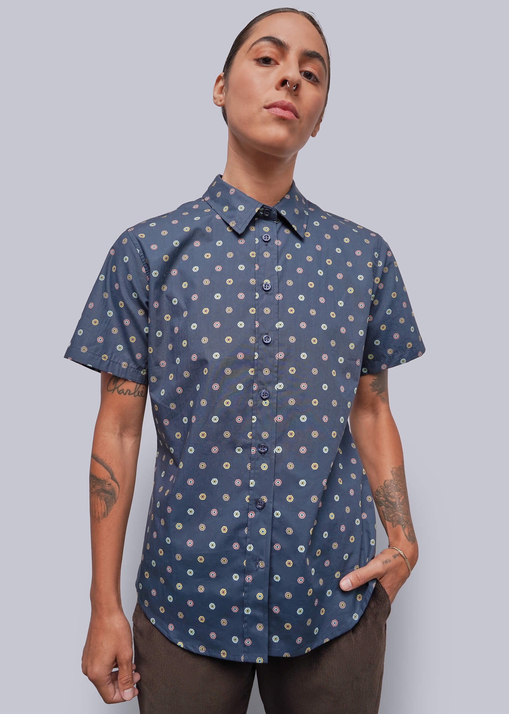 S=A model wearing a short-sleeve button-up shirt with a blue base and a small white and yellow floral pattern, paired with brown pants, against a solid gray background.
