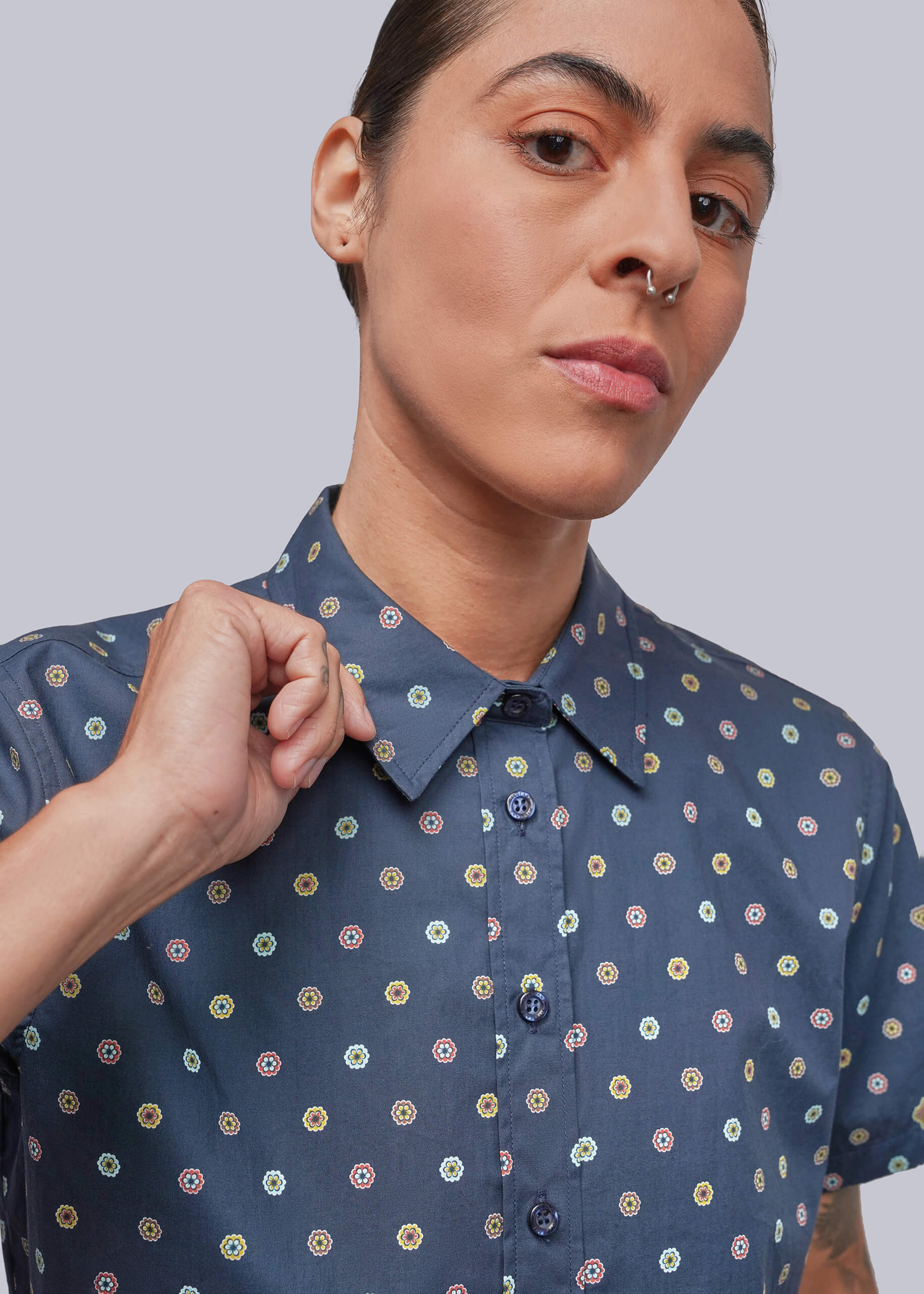 S=A close-up of a person wearing a short-sleeve button-up shirt in slate blue with a repeating multicolor circular pattern, set against a solid light gray background.