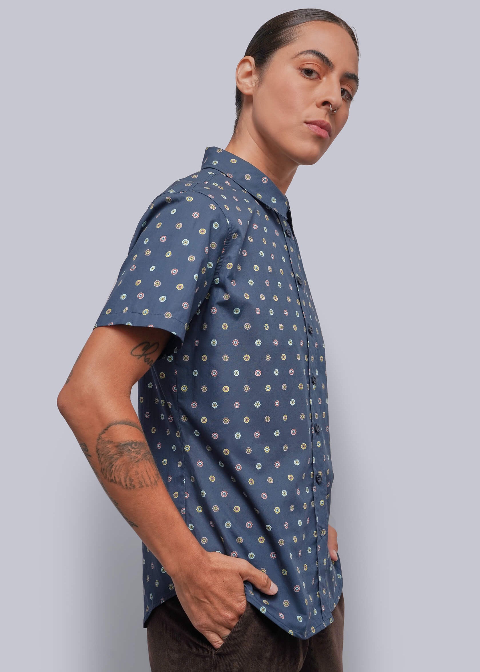 S=A person wearing a short-sleeve button-up shirt with a blue base and a small white and yellow geometric pattern, paired with dark brown pants, posed against a solid gray studio background.