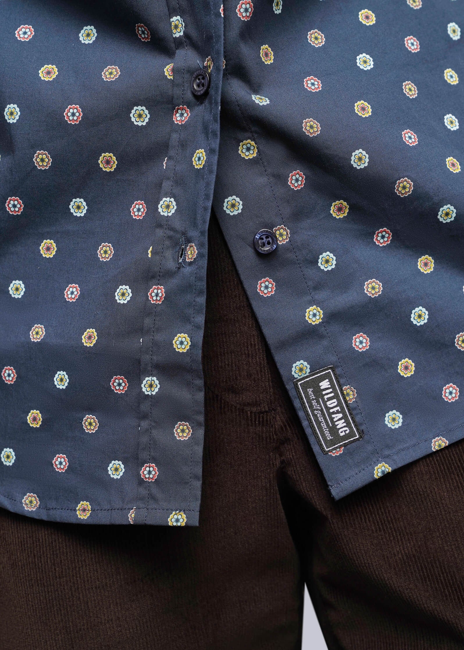 S=Close-up of a navy blue button-up shirt with a small multicolor geometric pattern, worn over dark brown corduroy trousers. A black brand label reading 'Mikrofashion' is visible near the hem.