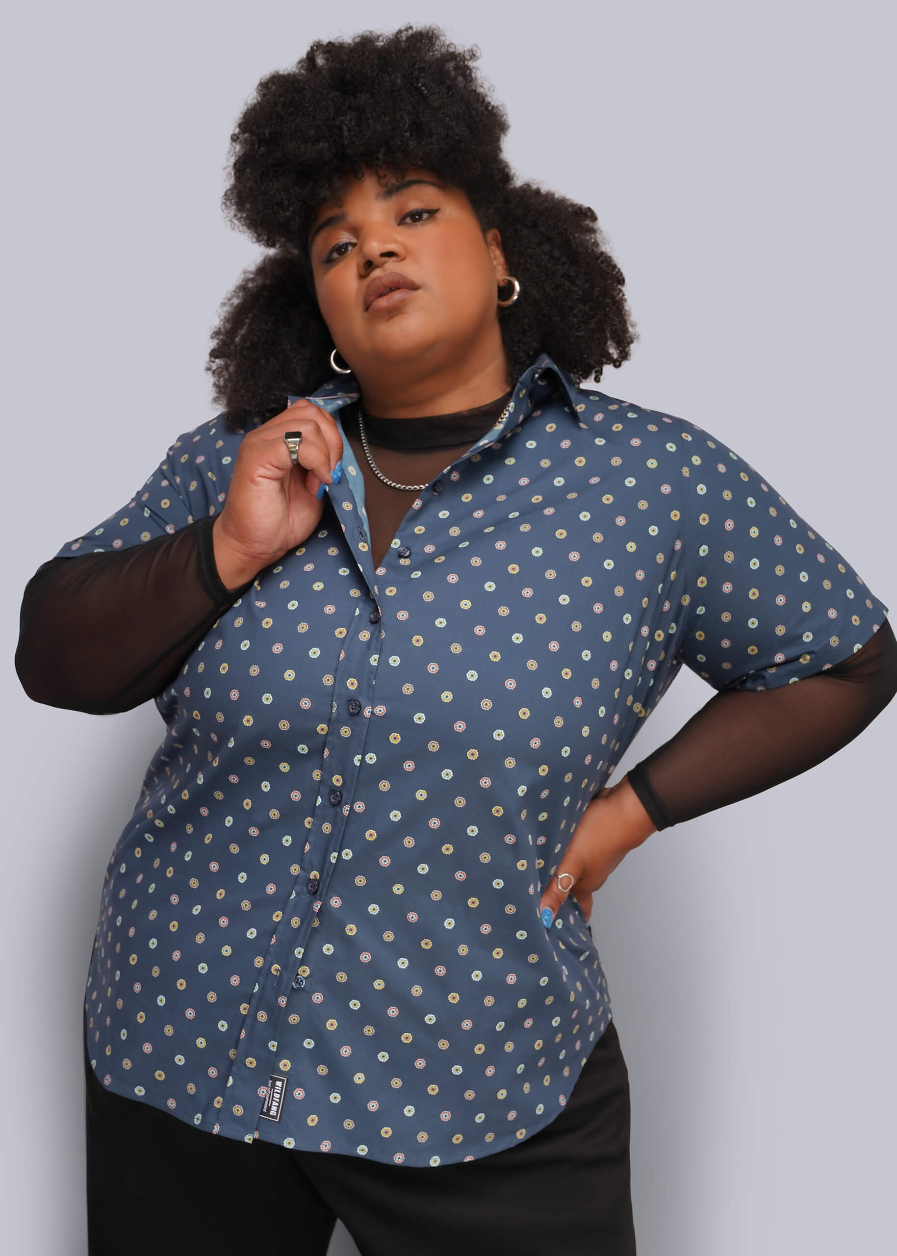 1X=A plus-size model wearing a blue short-sleeve button-up shirt with a white and yellow floral pattern, layered over sheer black long-sleeve sleeves, against a solid gray background.