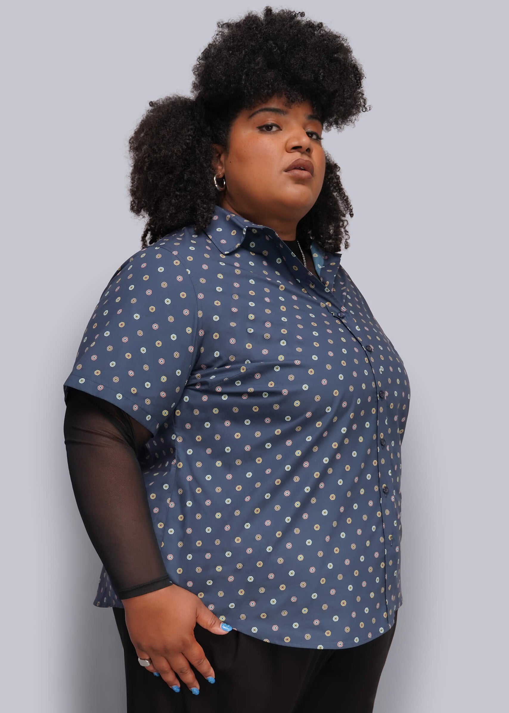 1X=A plus-size model wearing a short-sleeve button-up shirt in slate blue with a white and light blue polka dot pattern, layered over sheer black long-sleeve sleeves, against a solid light gray background.