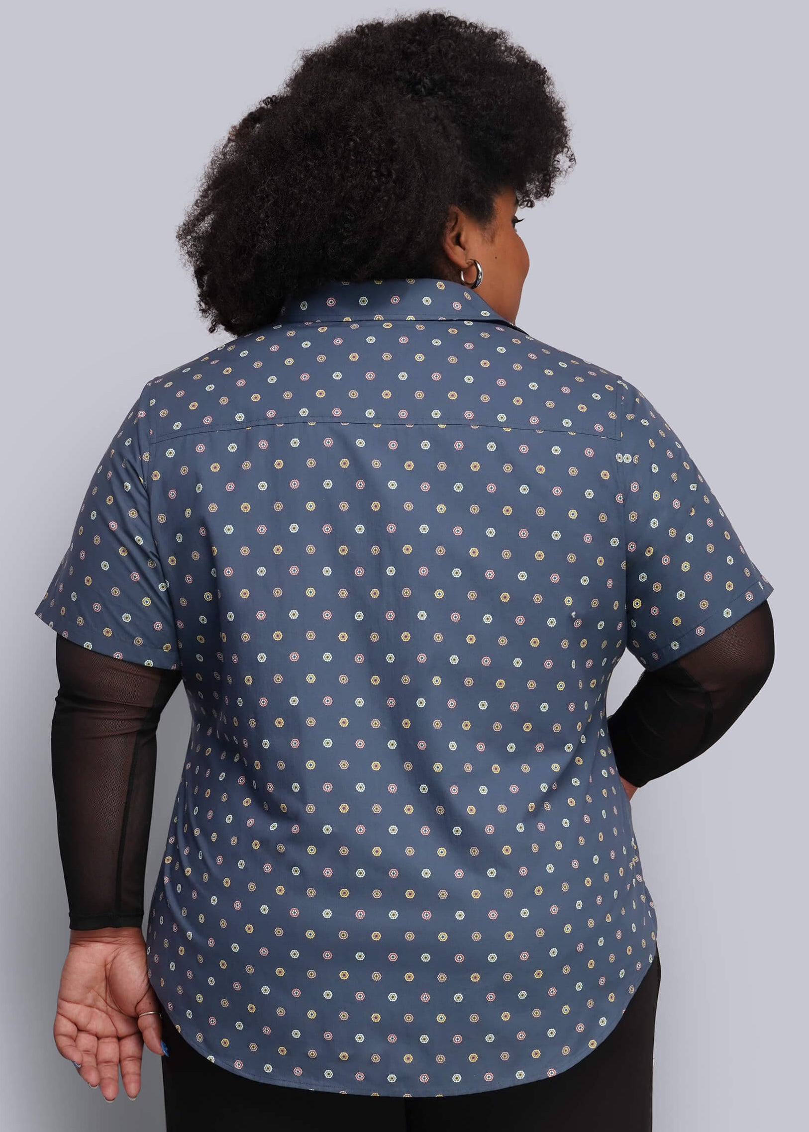 1X=A person shown from the back wearing a short-sleeve button-up shirt in blue with a small white geometric pattern, layered over black sheer long-sleeve sleeves and black pants, against a plain light gray studio background.