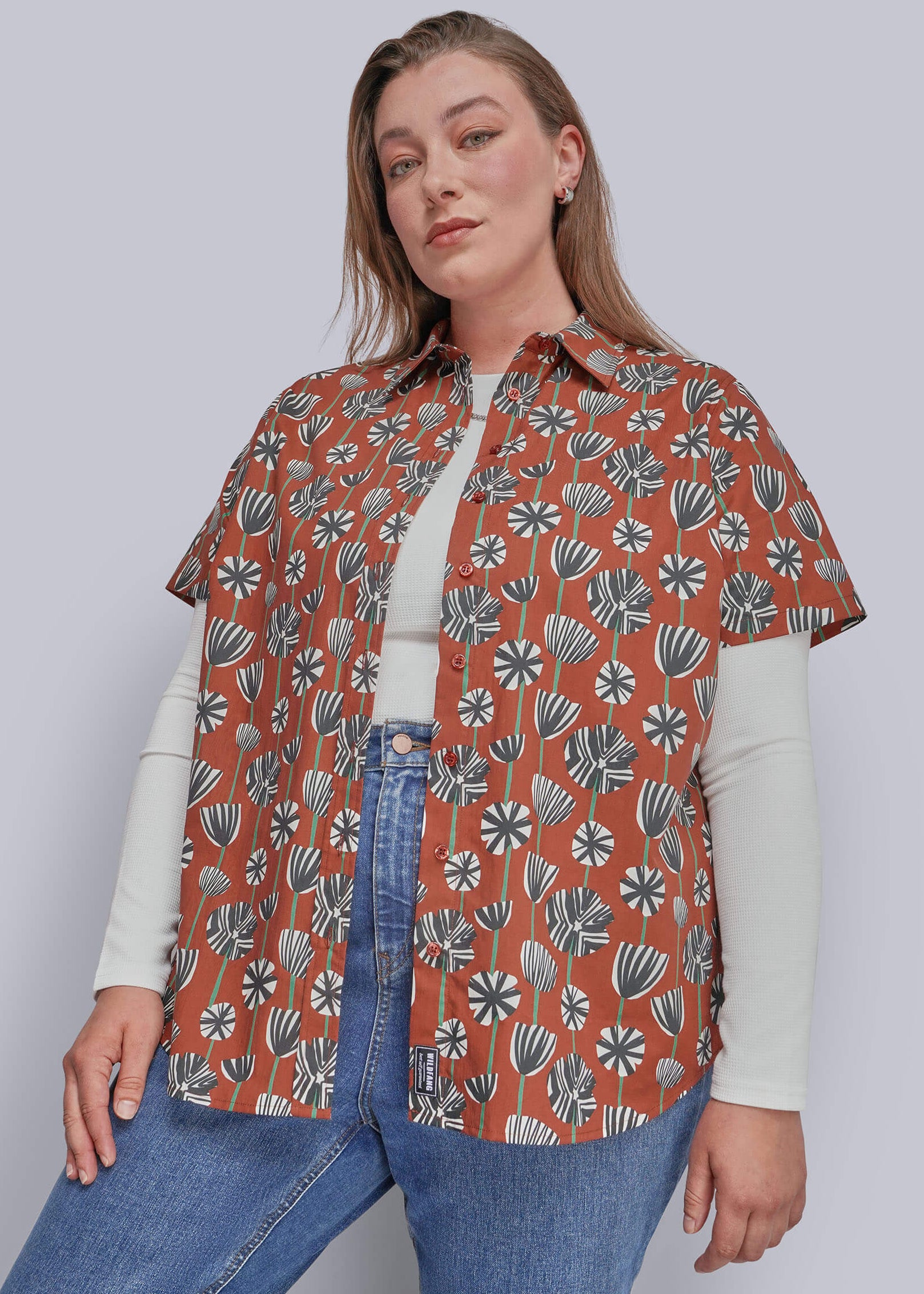 1X=A woman modeling a rust-orange short-sleeve button-up shirt with a white, gray, and green botanical pattern, layered over a white long-sleeve top and blue jeans against a solid gray studio background.