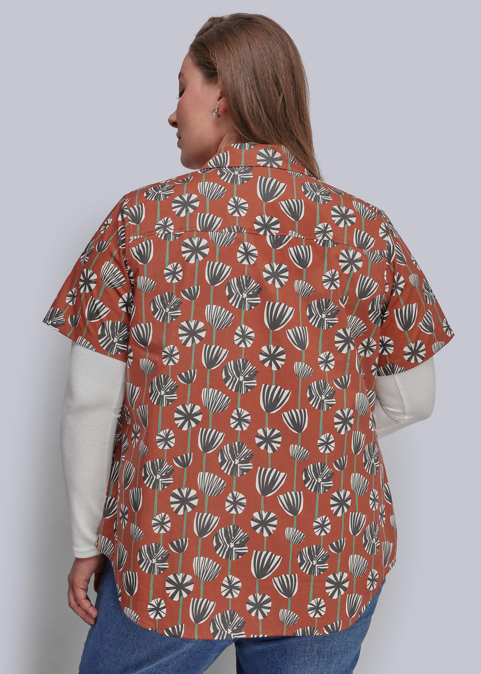 1X=A woman shown from the back wearing a rust-orange short-sleeve button-up shirt with a white and gray floral pattern, layered over a white long-sleeve top and blue jeans, against a solid gray studio background.