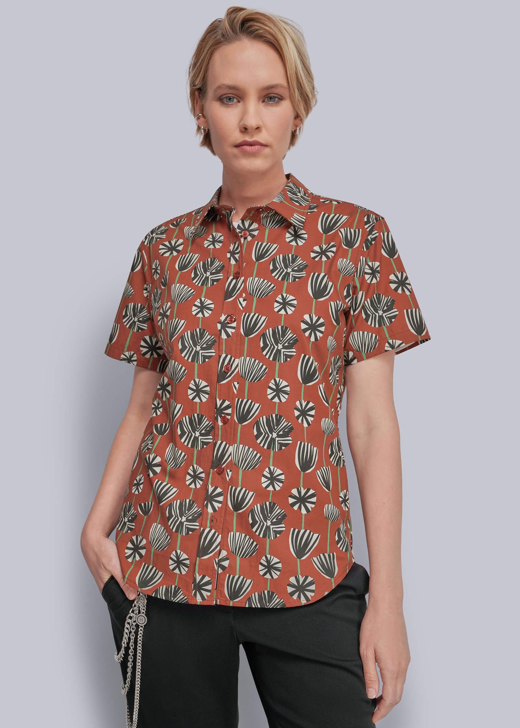 S=A woman modeling a short-sleeve button-up shirt with a rust-orange base and a black, gray, and white botanical pattern, paired with black pants against a solid gray studio background.