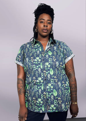 1X= Wearing The Essential Button Up, a tailored, navy blue shirt with green floral patterns and short sleeves, made of 100% cotton, they stand confidently against a plain backdrop. Dreadlocks frame their face, and tattoos are visible on their arms as they gaze at the camera.