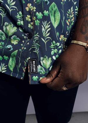 1X=A person wearing The Essential Button Up, a tailored fit, short-sleeved, 100% cotton shirt with a dark floral pattern and visible WILDFANG label, holds the hem. Their dark skin is accented by a gold ring and bracelet.