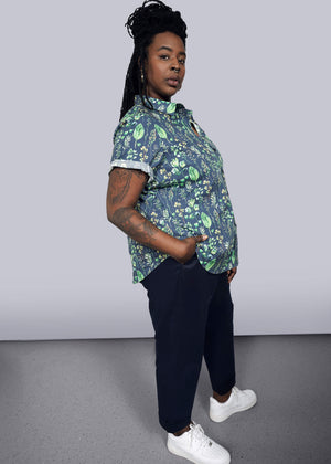 1X= With long braided hair, the person stands confidently with one hand in their pocket, wearing The Essential Button Up, a tailored fit leaf-patterned shirt with short sleeves, paired with dark pants and white sneakers. Made from 100% cotton, they look assuredly towards the camera.