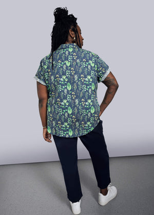 1X=Wearing The Essential Button Up, a short-sleeved, 100% cotton shirt in a tailored fit with a green leaf pattern, the person with long braided hair stands back to the camera. They pair it with dark pants and white sneakers against a plain, minimalistic background.