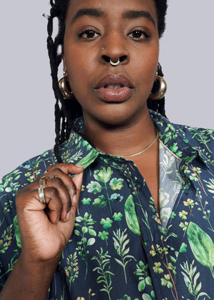 1X=A person confidently poses with hoop earrings and a septum ring, wearing The Essential Button Up—a tailored-fit, 100% cotton floral shirt with short sleeves—and holds its collar against a plain white background.