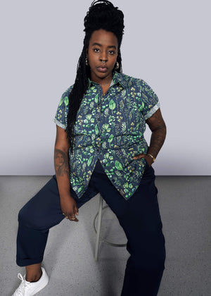 1X=A person with long braided hair sits on a stool against a gray background, wearing The Essential Button Up, a tailored fit green and navy floral short-sleeve shirt. They pair it with navy pants and white sneakers, with one hand in a pocket revealing tattoos on their arms.