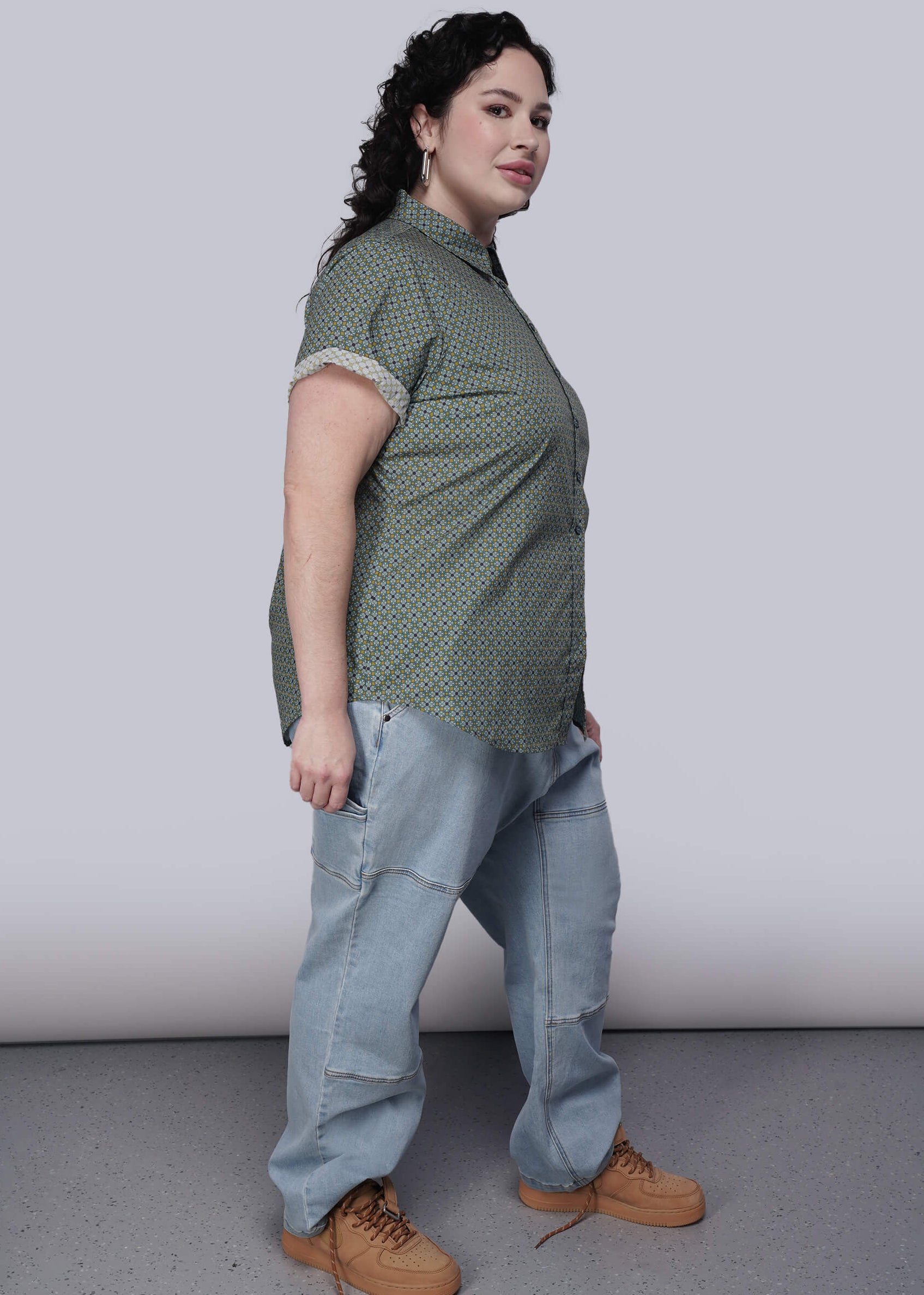 1X=Person wearing a green patterned shirt and light blue jeans with brown shoes on a plain background