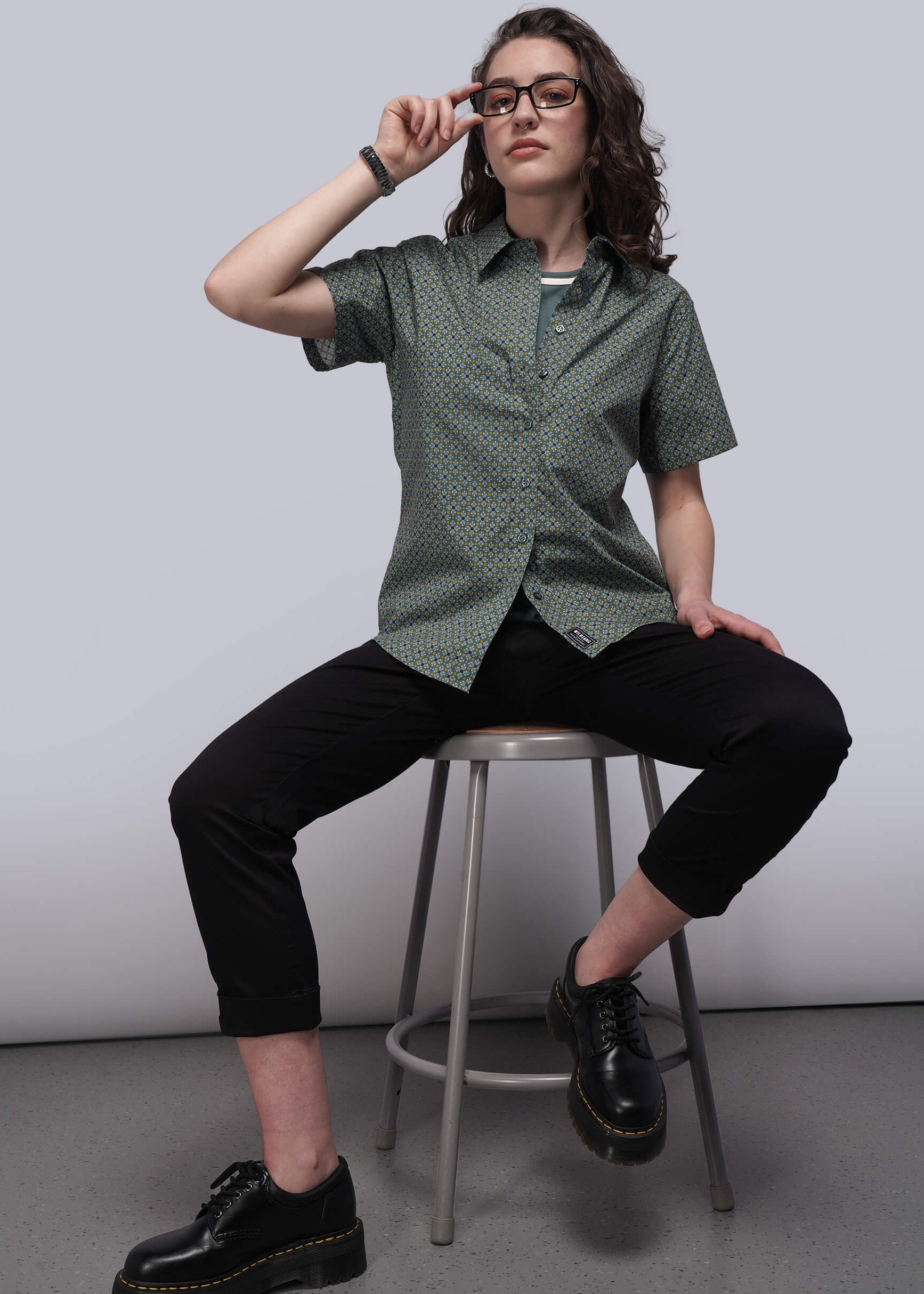 S=Person wearing a green patterned shirt and black pants sitting on a stool against a plain background
