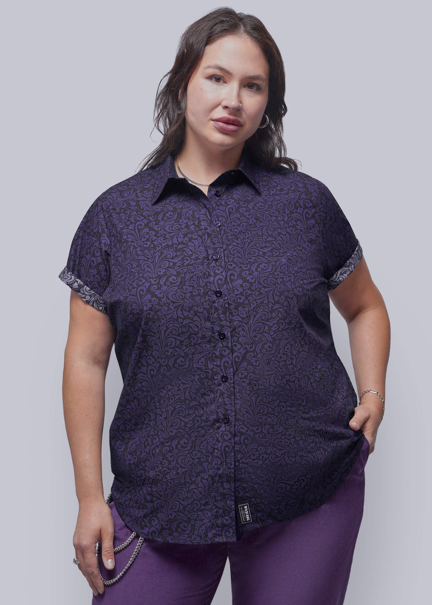 1X=A person modeling a short-sleeve button-up shirt with a dark purple and black abstract pattern, paired with purple pants, against a solid gray studio background.
