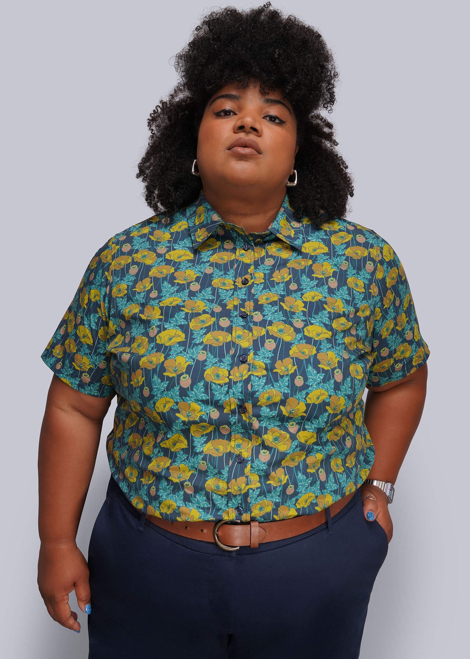 QX=A plus-size model wearing a short-sleeve button-up shirt with a yellow and teal floral pattern, paired with navy blue pants and a brown belt, against a solid gray background.