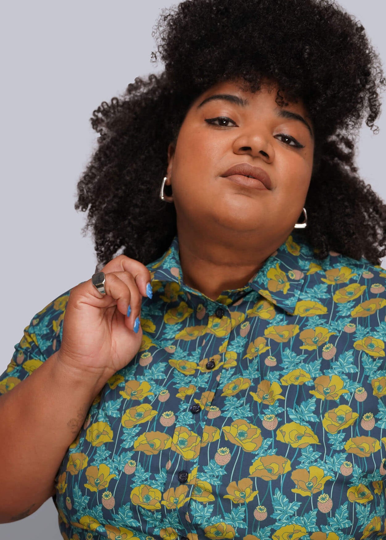 1X=Close up of model wearing a short-sleeve button-up shirt with a yellow and teal poppy floral pattern.