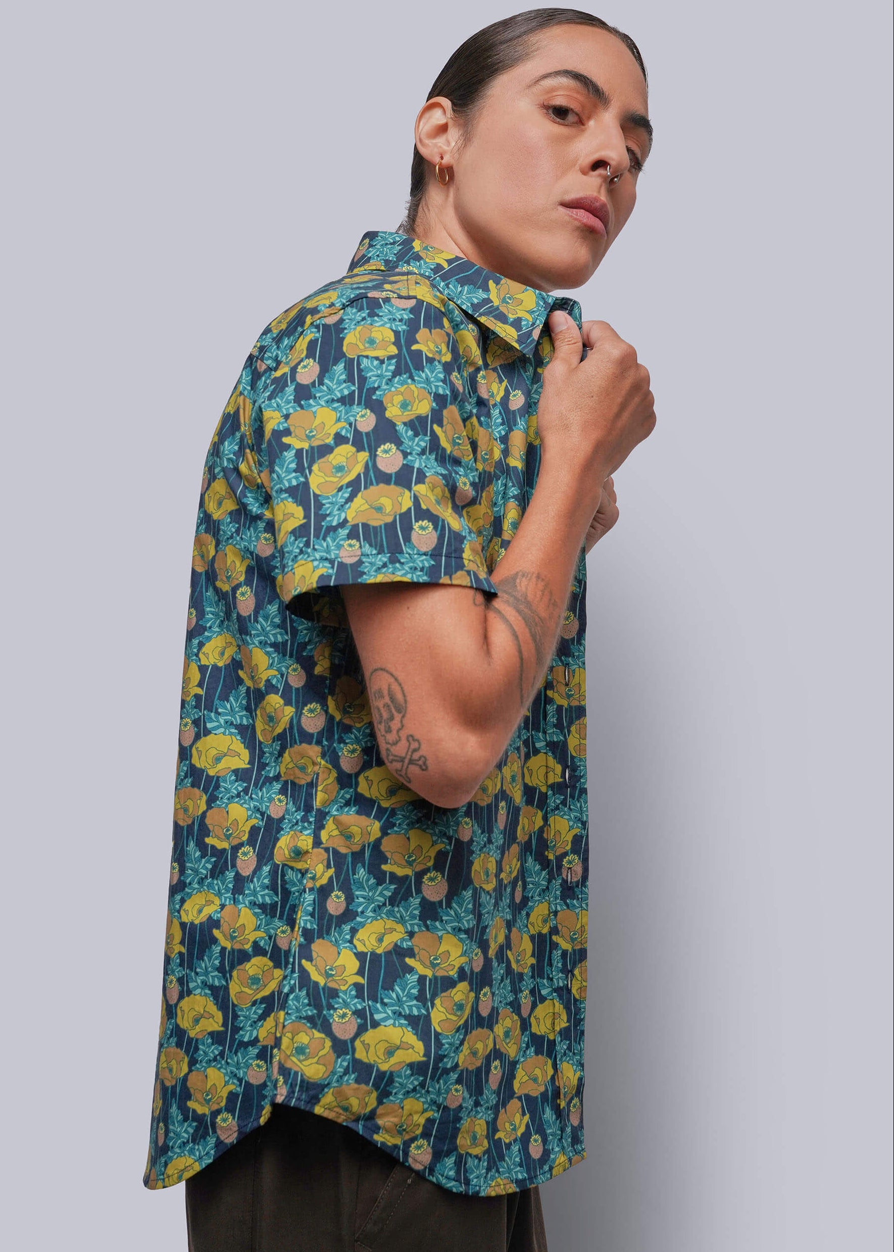 S=A person wearing a short-sleeve button-up shirt with a yellow and blue floral pattern, posed against a solid gray background.