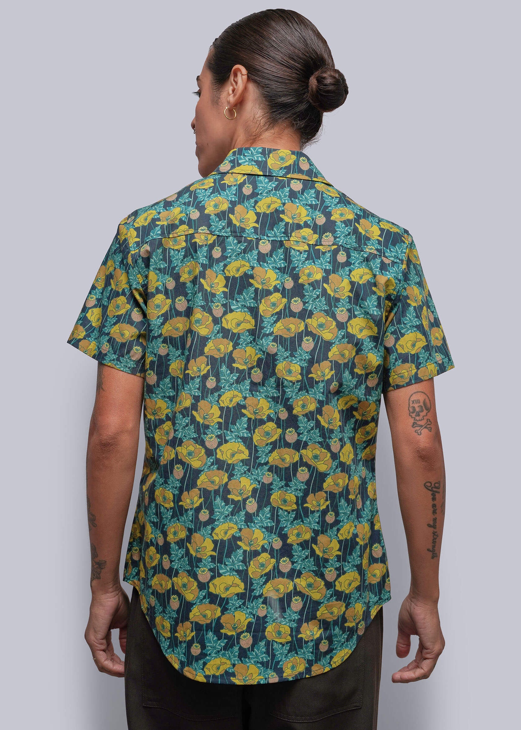 S=A person viewed from the back wearing a short-sleeve button-up shirt with a dense yellow and teal floral pattern on a dark background, paired with dark trousers, set against a solid gray studio backdrop.