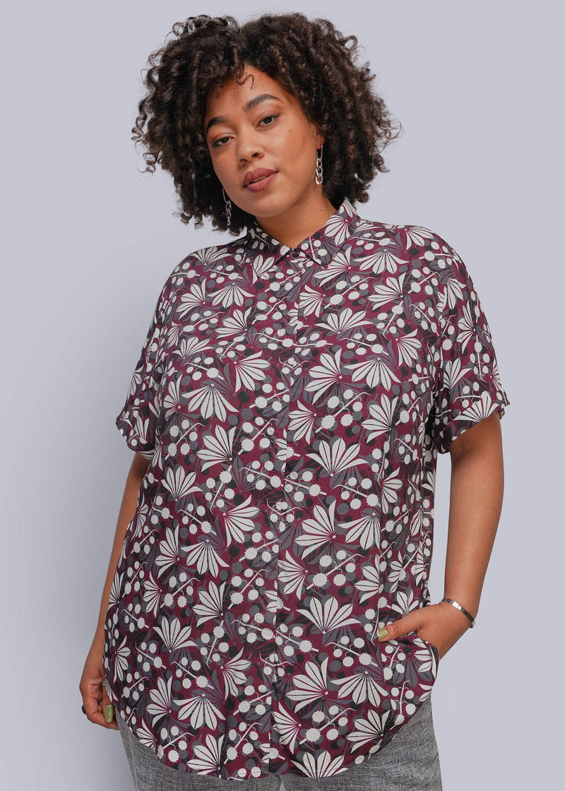 1X=A woman with curly dark hair models a short-sleeve button-up shirt featuring a burgundy, white, and gray floral pattern, paired with gray pants, against a solid light gray background.