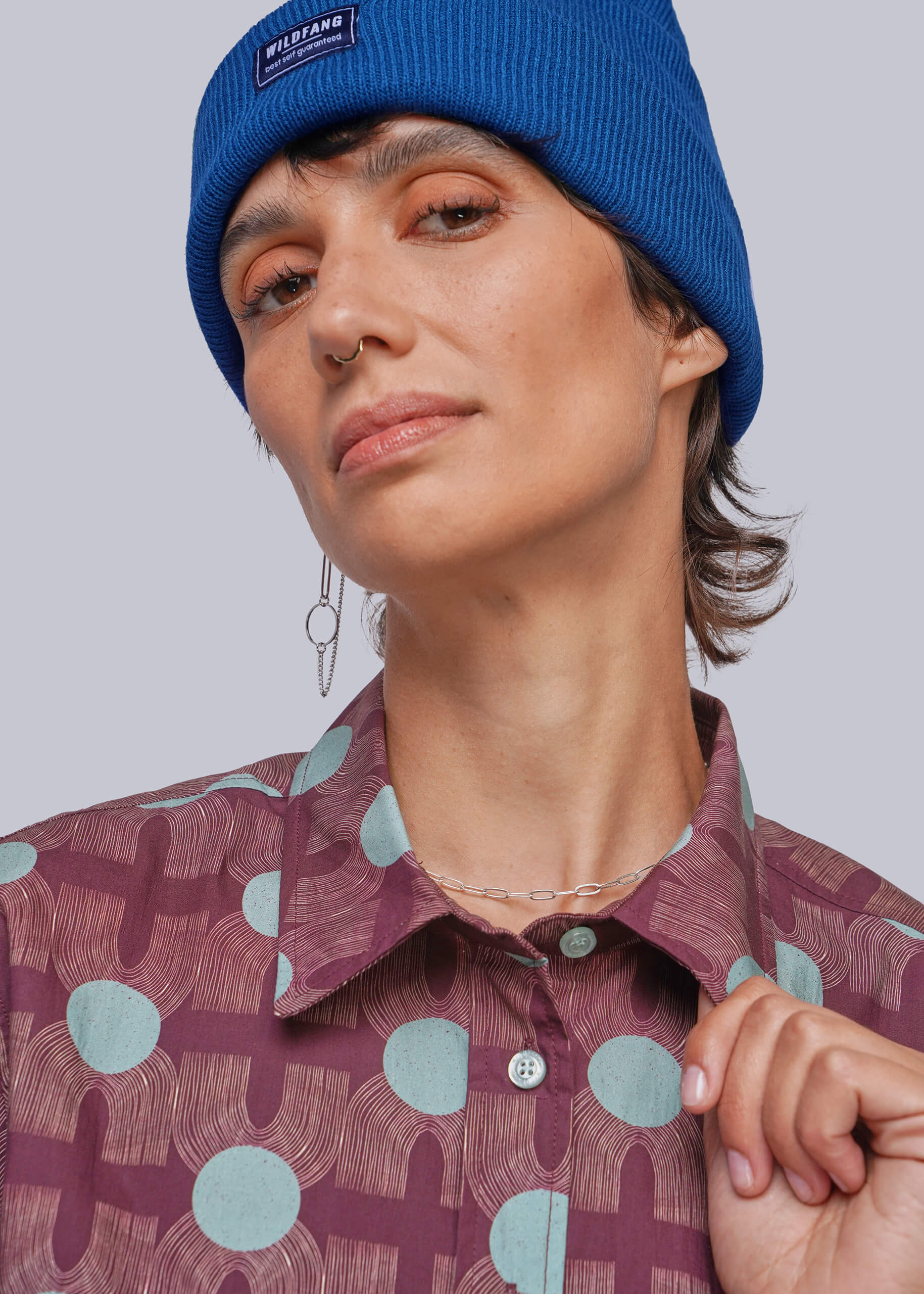 A close-up of a person wearing a bright blue ribbed knit beanie with a small black brand patch that reads 'WILDFANG' and a patterned button-up shirt in mauve and light blue.