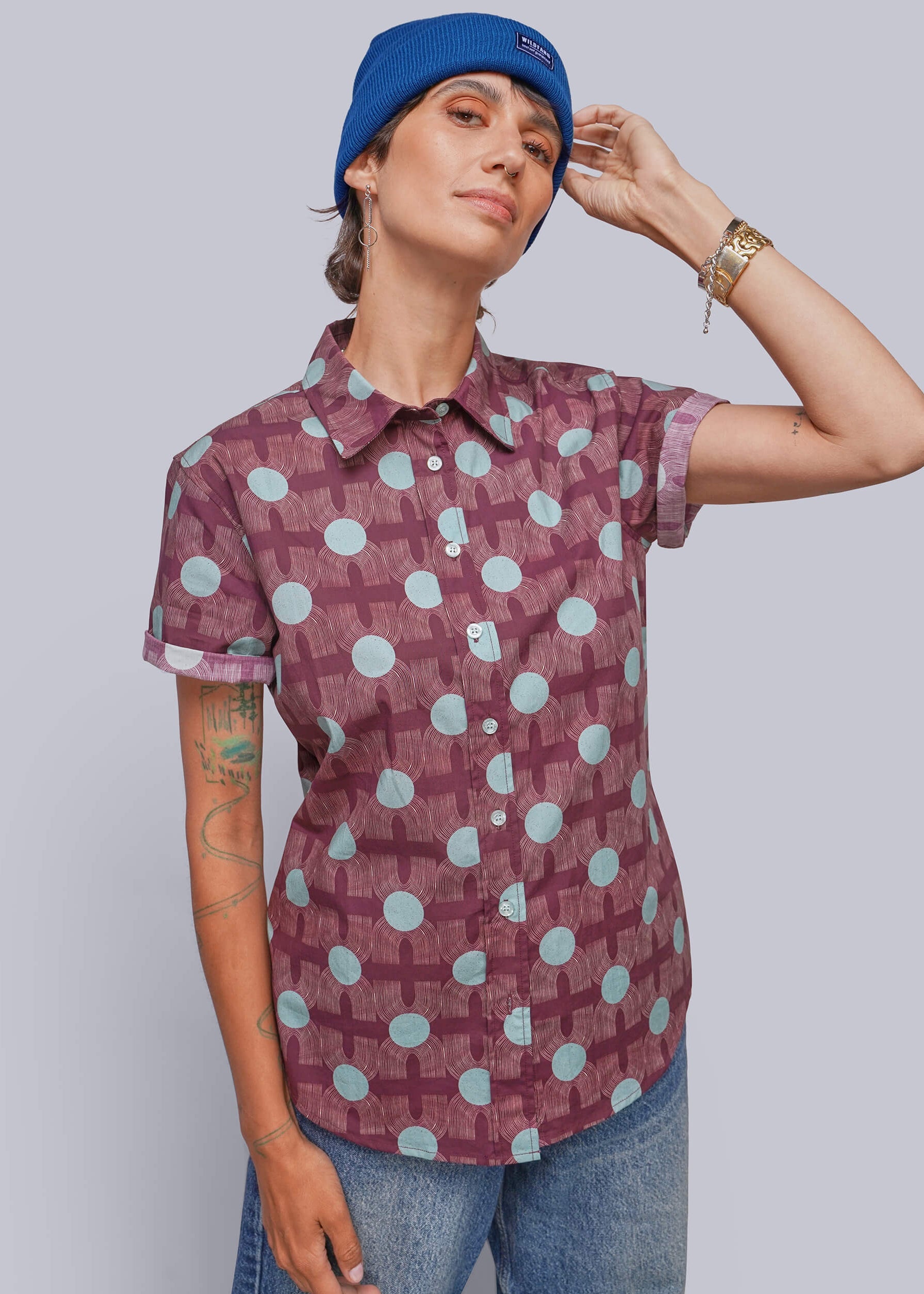 A person wearing a short-sleeve button-up shirt with a maroon and light blue geometric pattern, paired with blue jeans and a blue beanie, posed against a solid gray studio background.