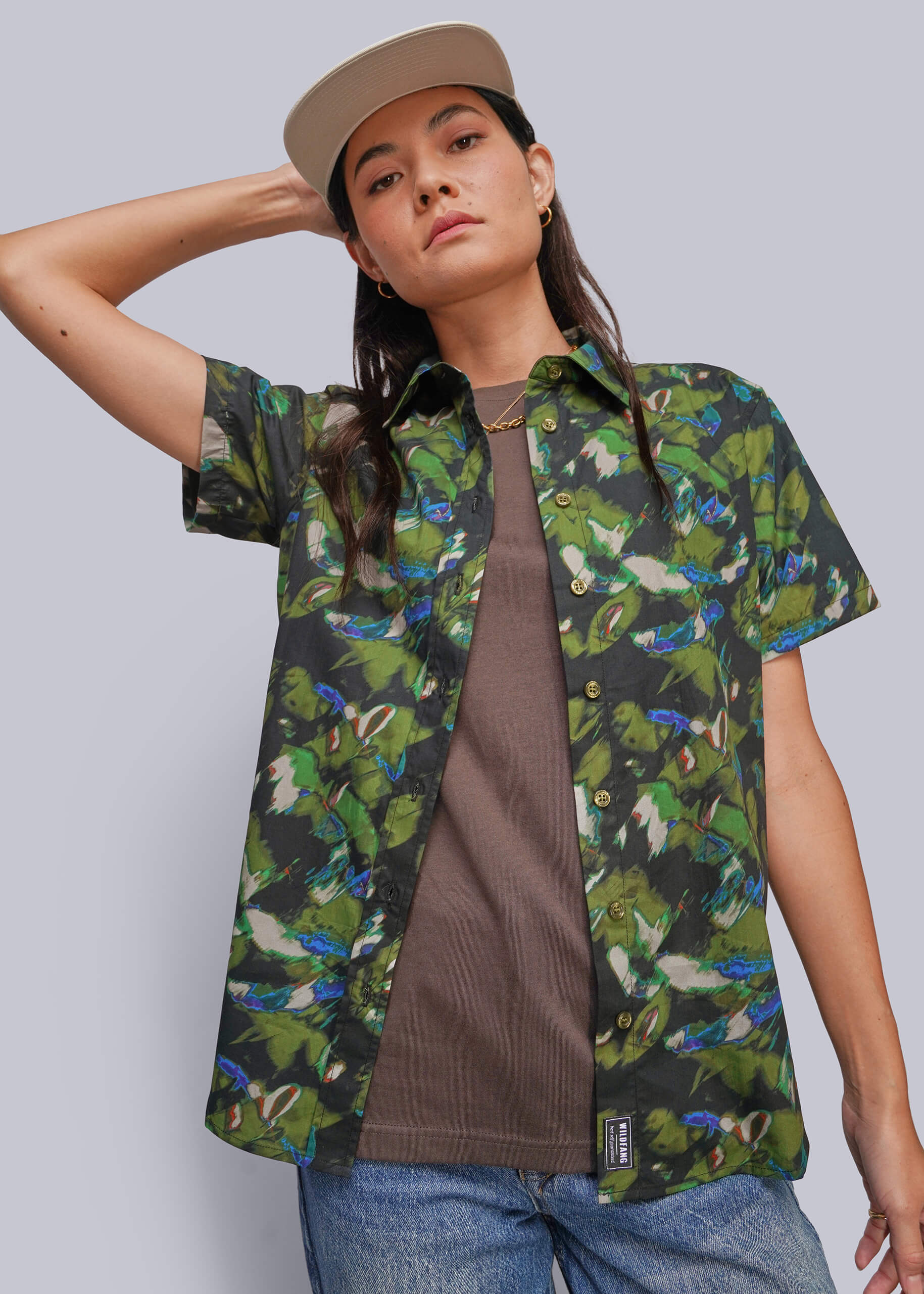 A model wearing a green tropical parrot print short-sleeve button-up shirt over a brown t-shirt, blue jeans, and a beige cap, set against a solid gray studio background.