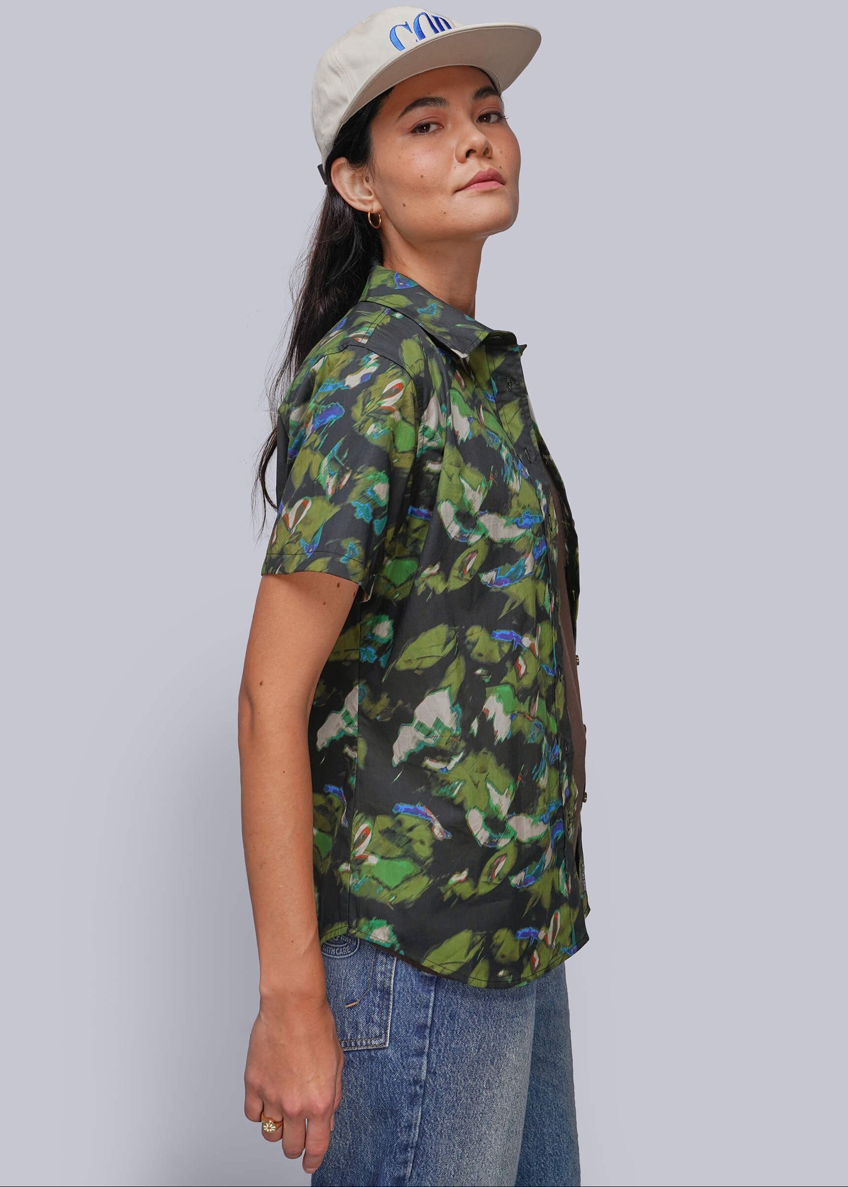 A woman wearing a short-sleeve button-up shirt with a green and black tropical leaf pattern, paired with blue jeans and a beige baseball cap, shown in a side-profile view against a solid gray background.