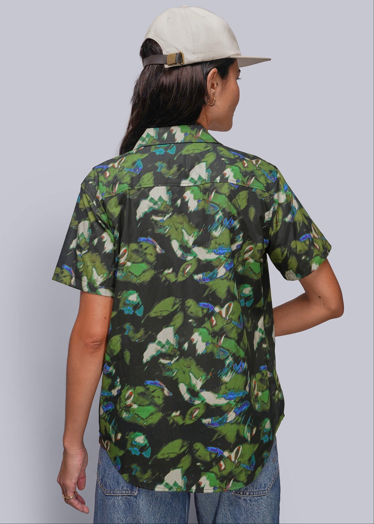 S=A person shown from the back wearing a short-sleeve button-up shirt with a green camouflage-style leaf print on a black background, paired with a light beige cap and blue jeans, against a plain light gray studio background.