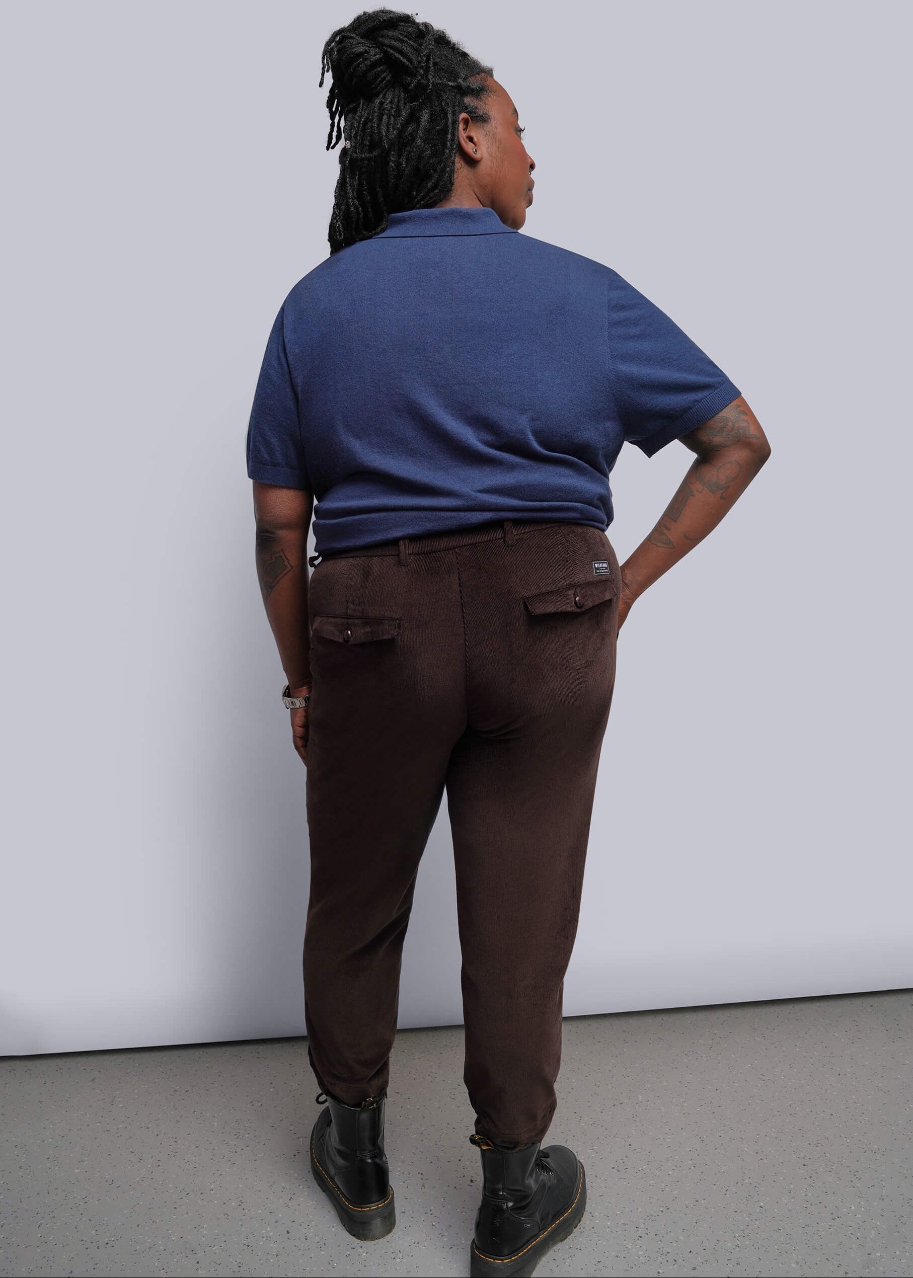 1X=A person shown from the back wearing a blue short-sleeve polo shirt and dark brown trousers, standing against a plain light gray wall.