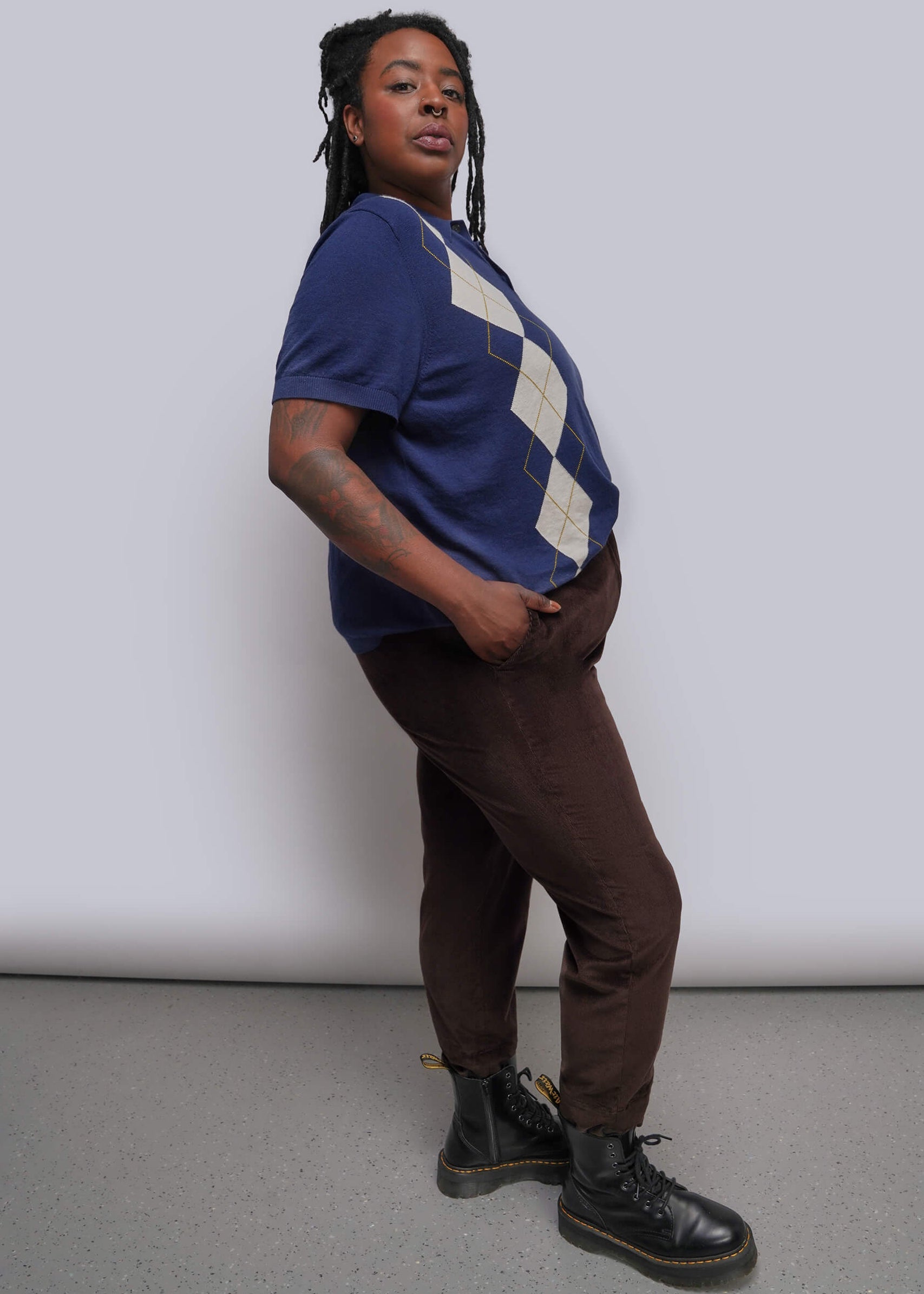 1X=A person modeling a short-sleeve sweater with a navy blue and beige argyle pattern, paired with dark brown pants and black boots, standing against a light gray studio backdrop.