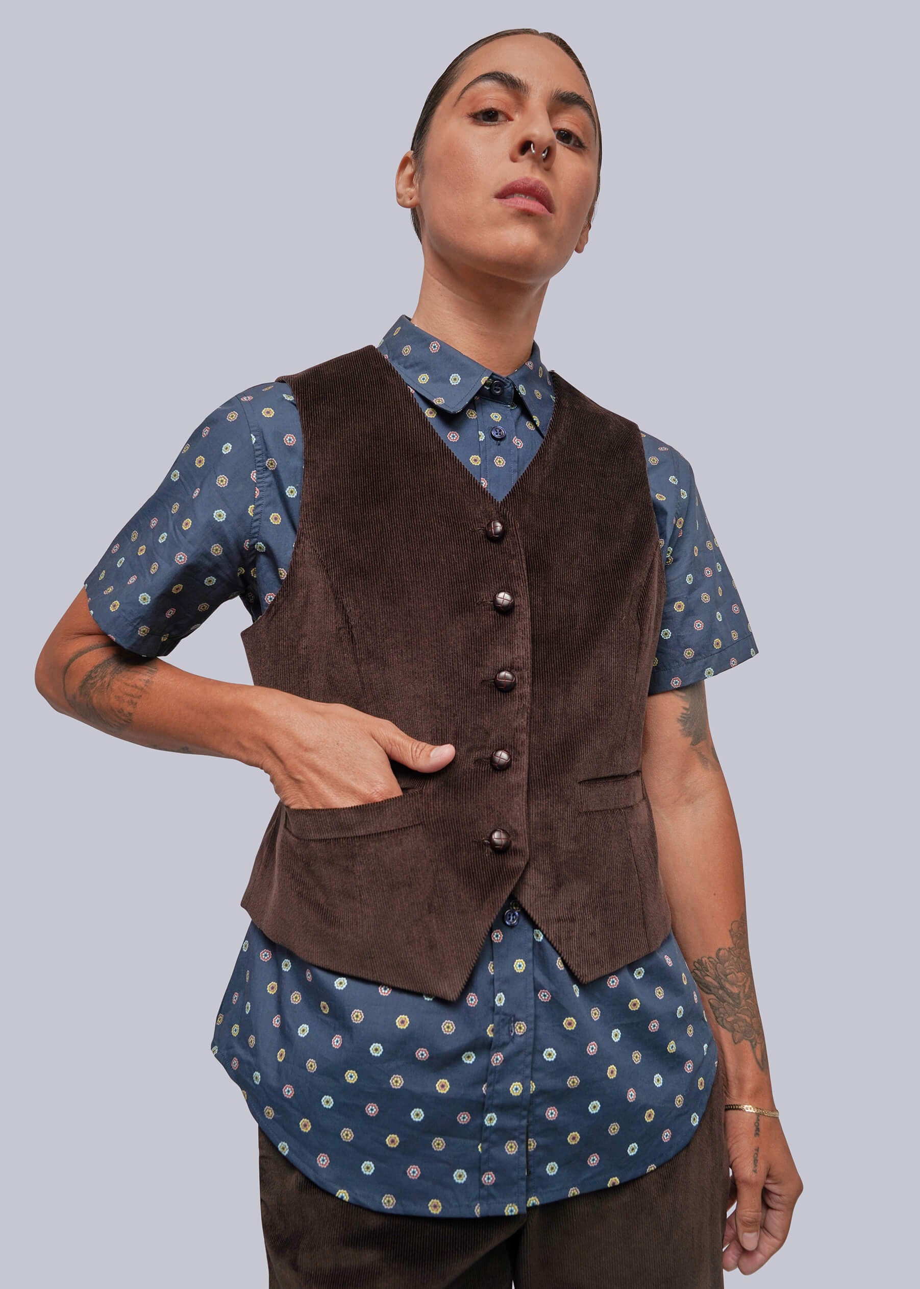 A model wearing a dark brown corduroy vest over a blue and white patterned short-sleeve button-up shirt, paired with dark brown corduroy trousers, against a solid gray background.