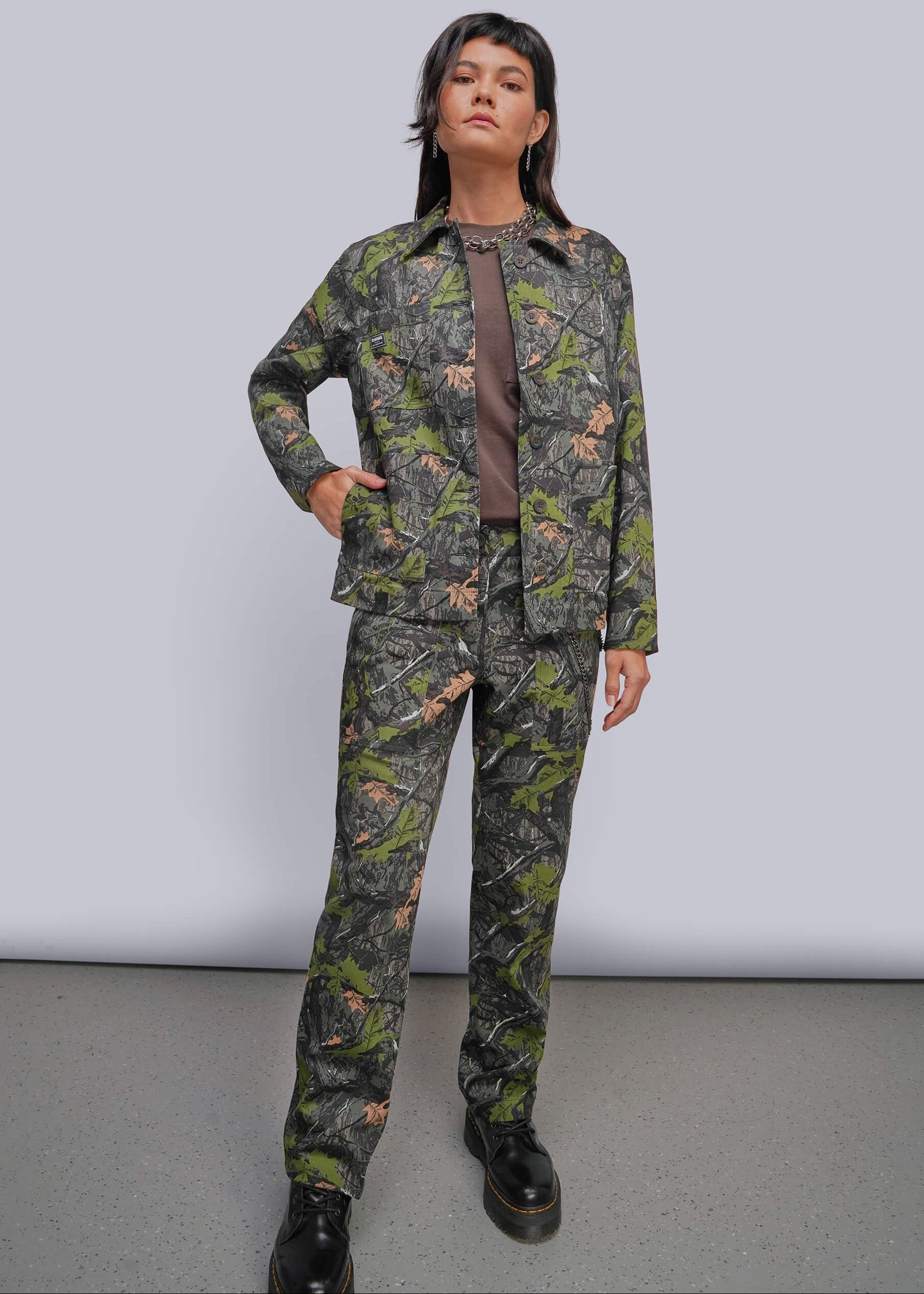 A model stands in a studio wearing a matching camouflage jacket and pants set with a brown shirt underneath and black boots. The camouflage pattern features green, brown, and black tones.