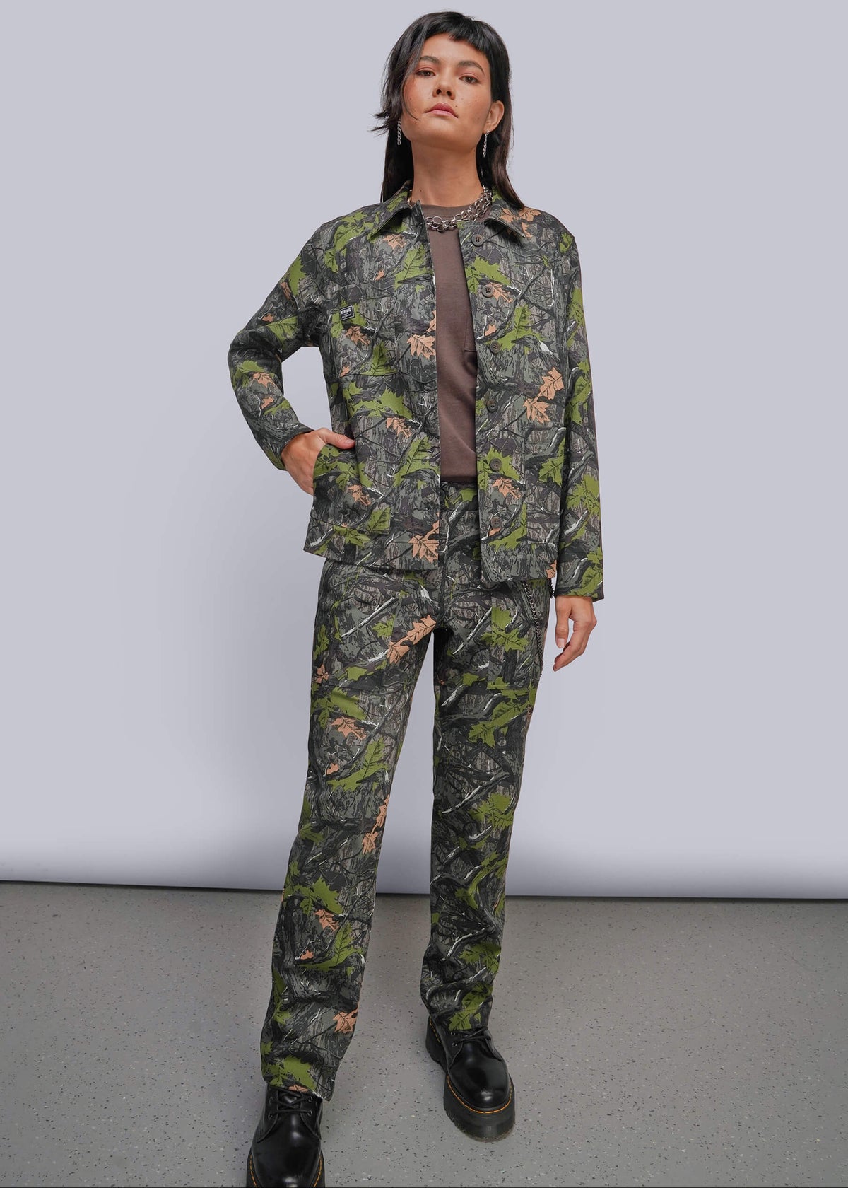S=A model stands in a studio wearing a matching camouflage jacket and pants set with a brown shirt underneath and black boots. The camouflage pattern features green, brown, and black tones.