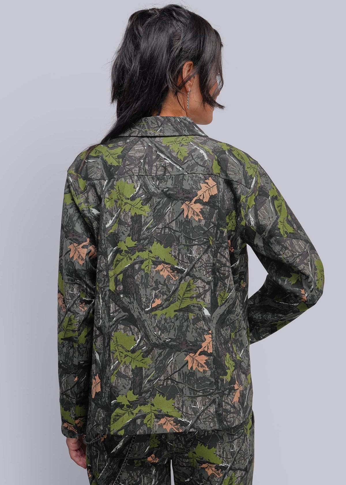 S=A model shown from the back wearing a long-sleeve jacket and matching pants in a dark green, brown, and black woodland camouflage pattern with brown leaf accents, set against a solid light gray background.