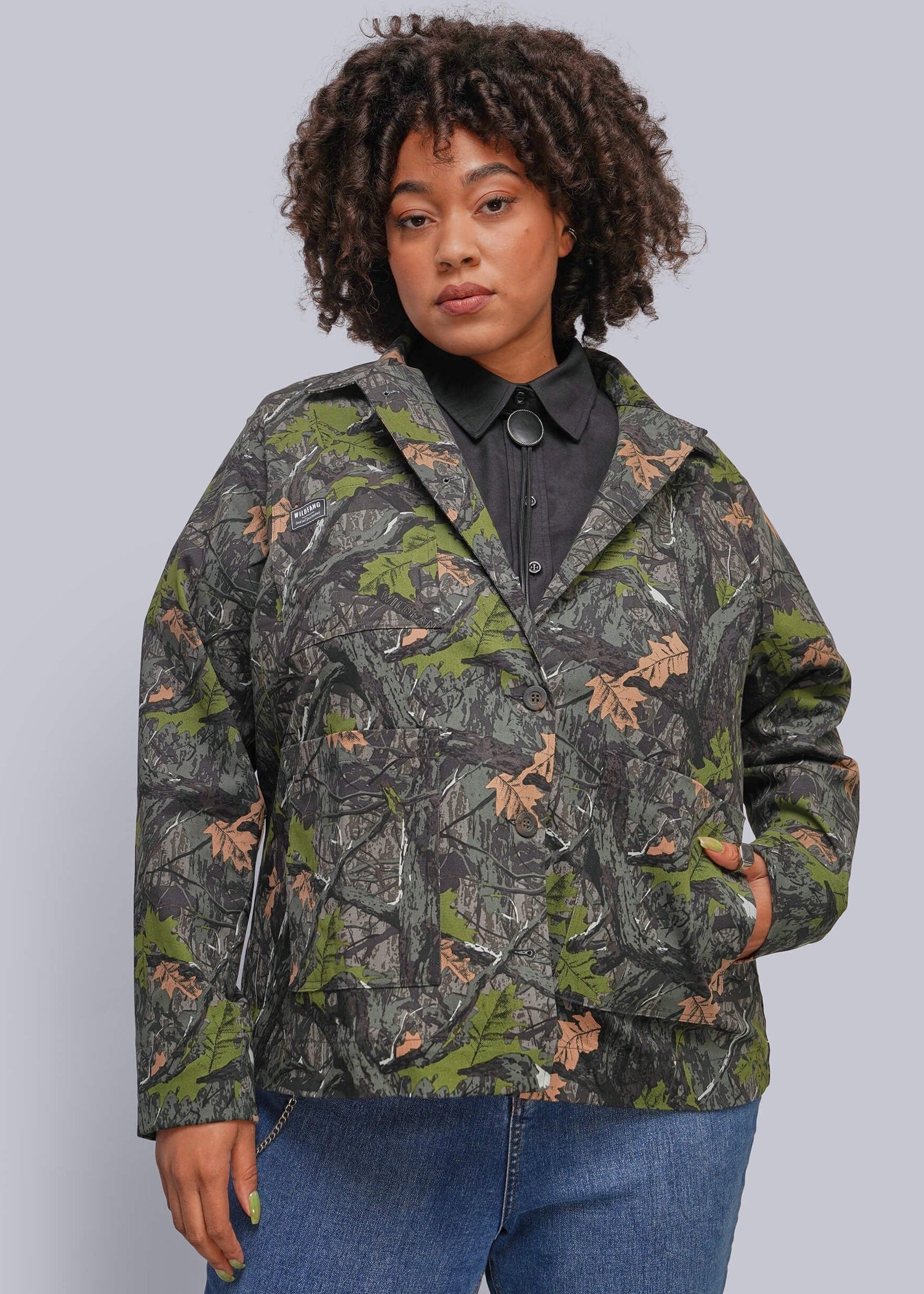 1X=A woman wearing a camouflage jacket with green, brown, and gray tones, styled over a black collared shirt and blue jeans, against a solid gray background.