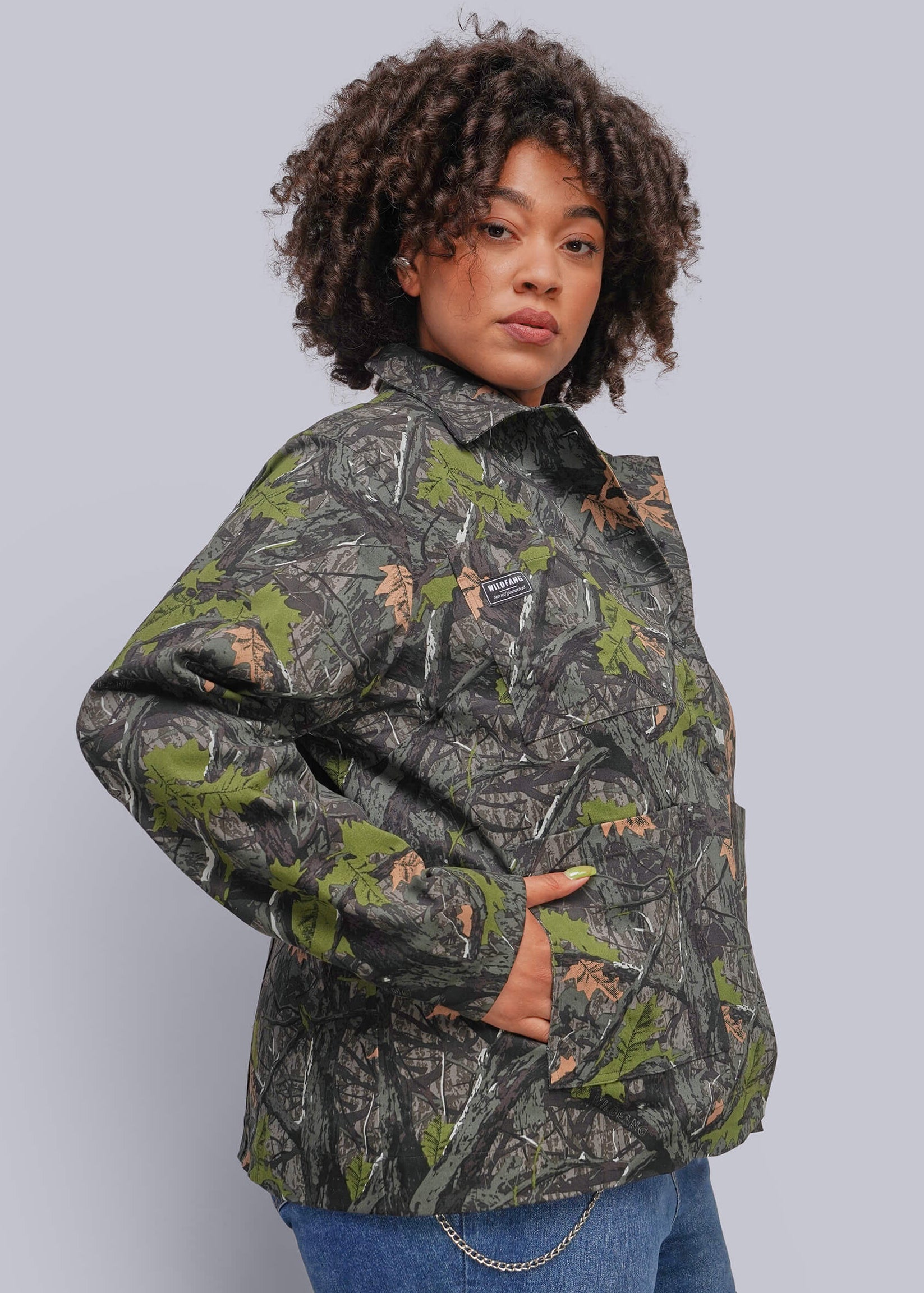 1X=A woman with curly dark hair models a camouflage jacket featuring green, brown, and gray woodland patterns, paired with blue jeans against a solid gray studio background.
