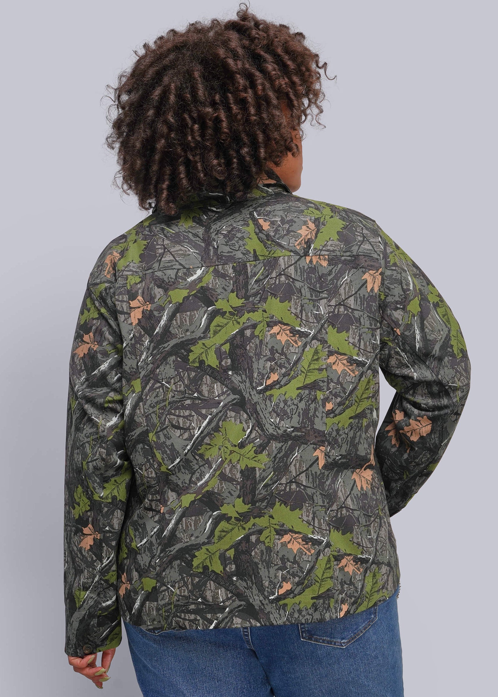 1X=A person shown from the back wearing a camouflage jacket with green, gray, and brown woodland print, paired with blue jeans, against a solid gray studio background.