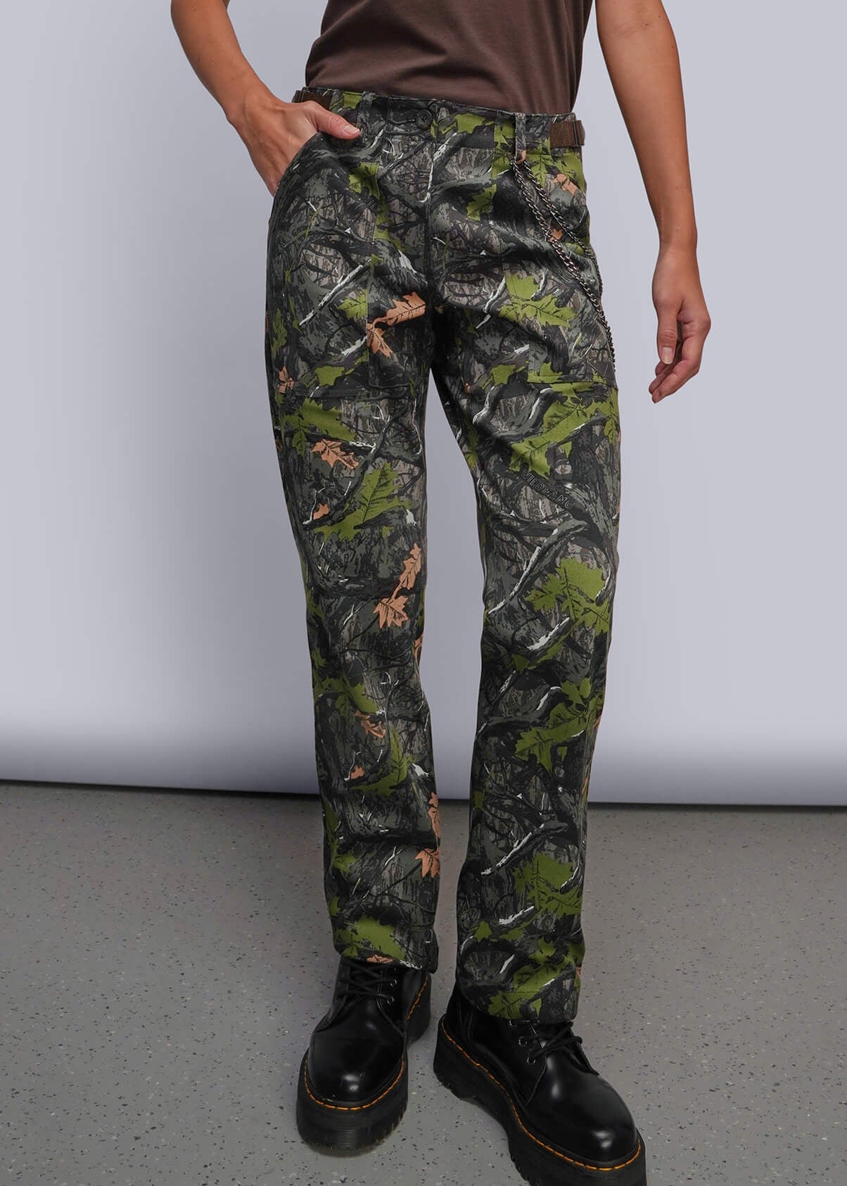 A model stands against a light gray studio background wearing a brown short-sleeve t-shirt, camouflage pants, and black boots.