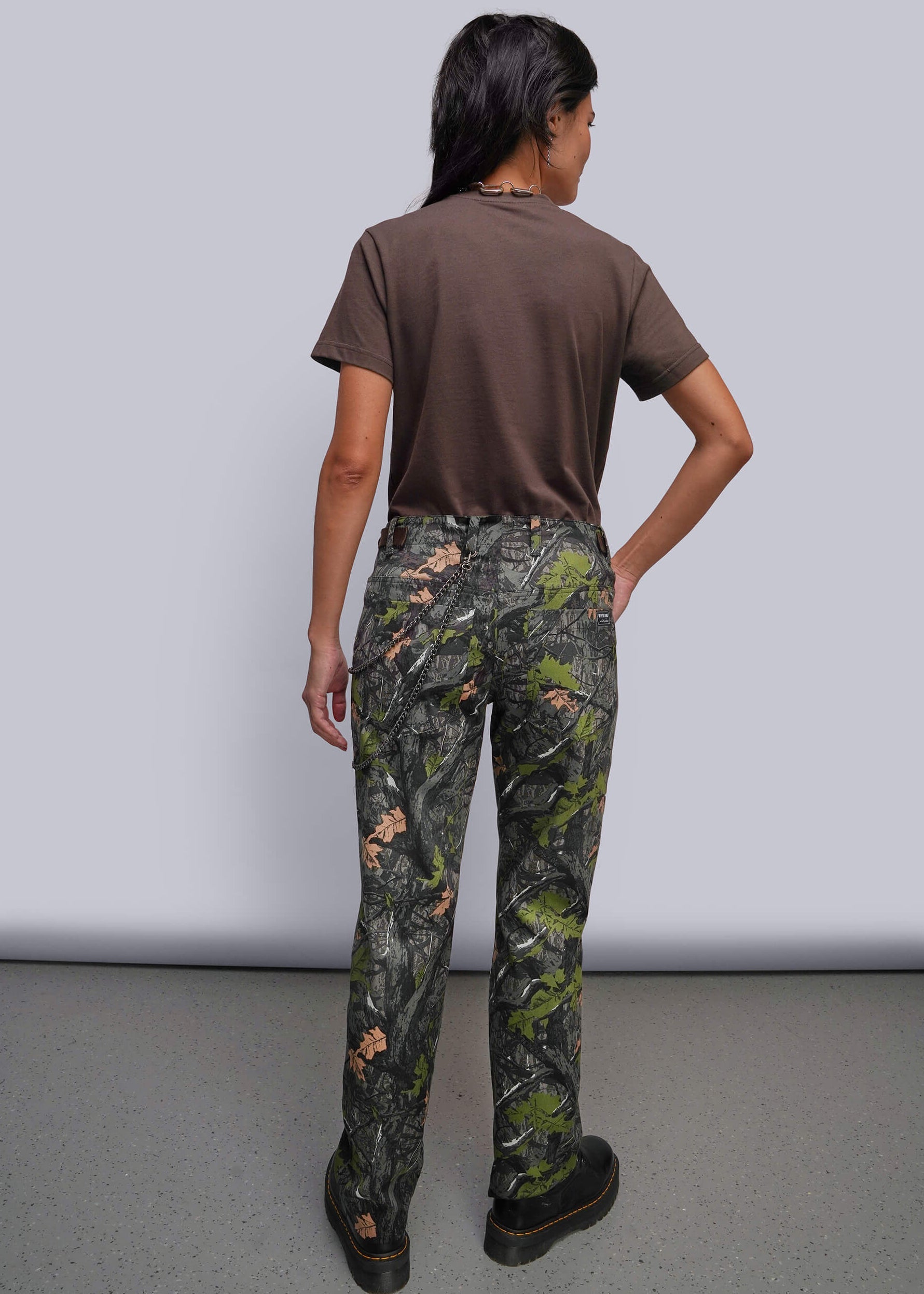 S=A model shown from the back wearing camouflage pants, a brown t-shirt, and black shoes against a plain gray studio background.